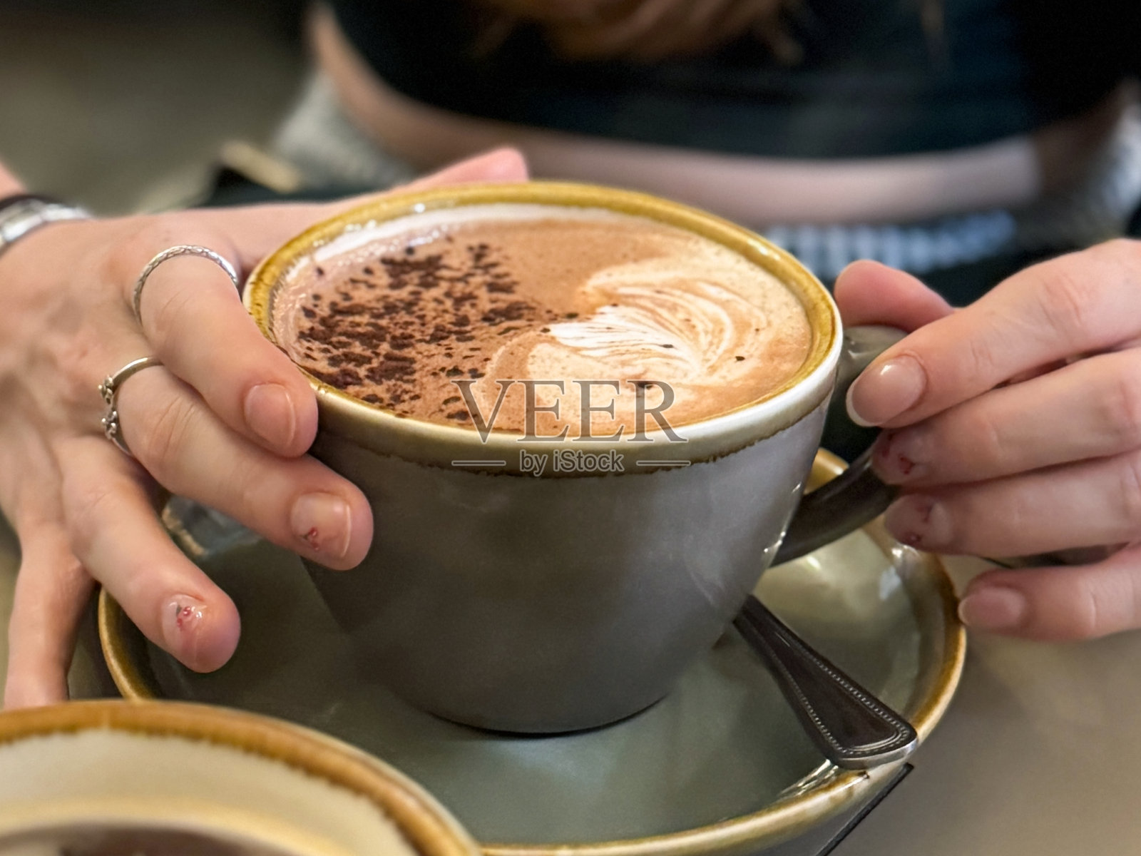 特写镜头，无法识别的女性手持一杯热巧克力，杯子为大号绿色，放在碟子上，表面撒有可可粉，展示了指甲油美甲设计，樱桃图案法式指甲艺术，咖啡馆环境，俯视角度，重点在前景。照片摄影图片