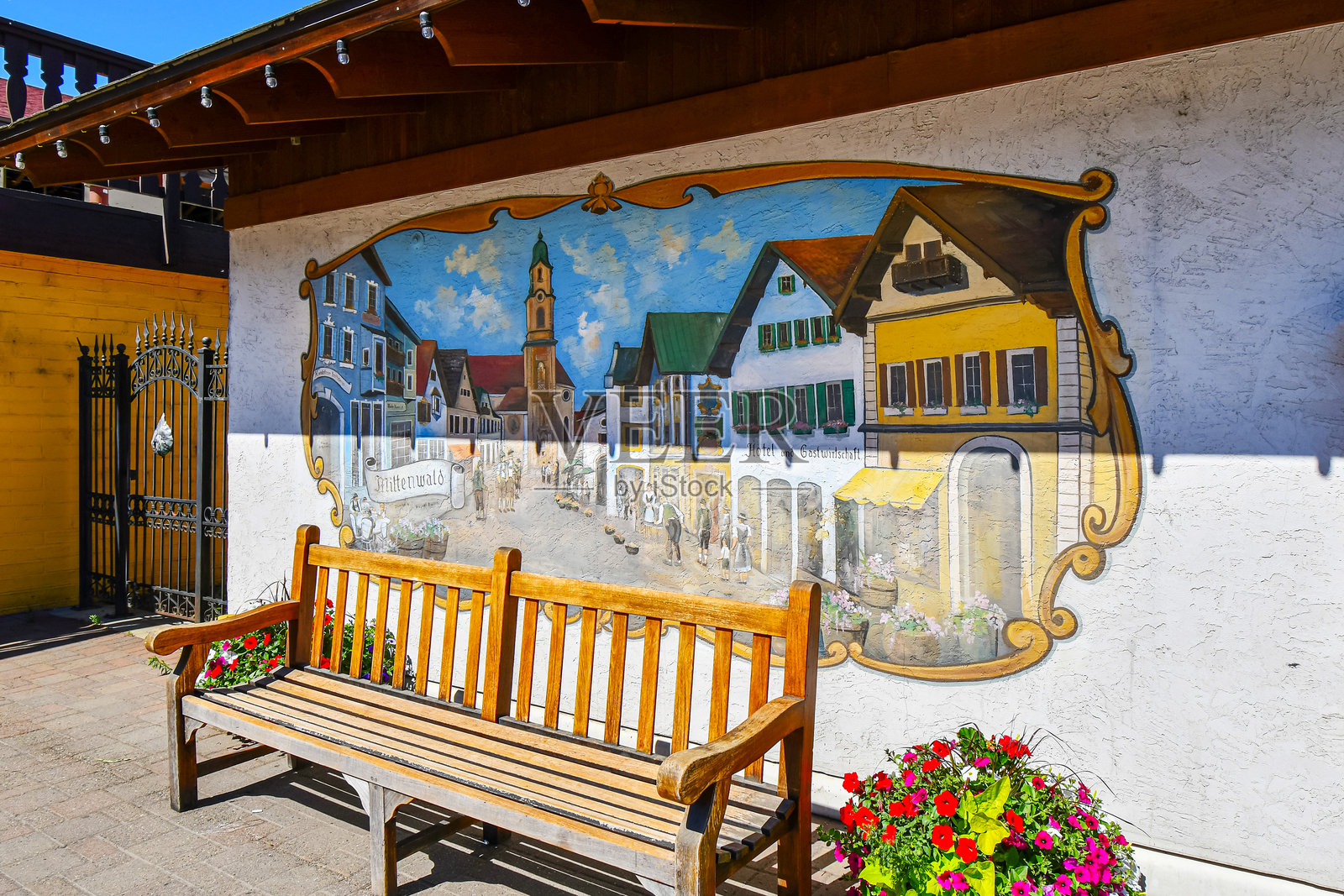 一张公园长椅旁边有花，前面是德国米滕瓦尔德镇的壁画，位于华盛顿州以巴伐利亚为主题的莱文沃斯镇。照片摄影图片