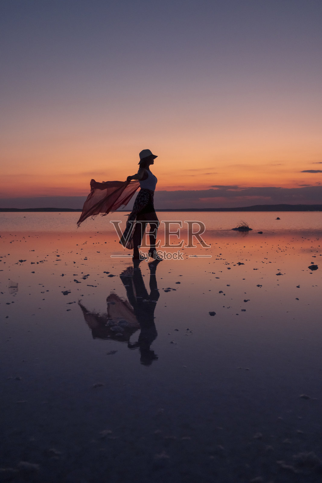 女人的剪影在日落时分走在宁静的盐湖上，手中挥舞着一条飘逸的围巾照片摄影图片