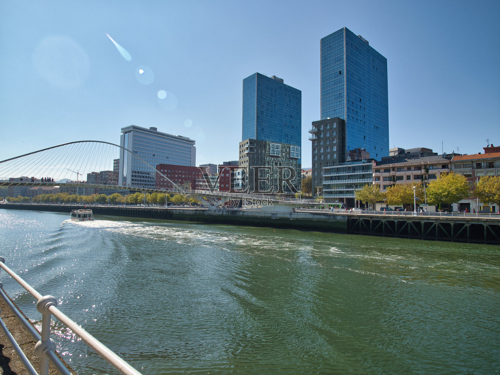 比尔巴鄂河畔城市景观与现代塔楼。- 西班牙比尔巴鄂的现代天际线，塔楼在宁尔维翁河中倒影，映衬在晴朗的蓝天之下，展示了城市发展和建筑风格。照片摄影图片