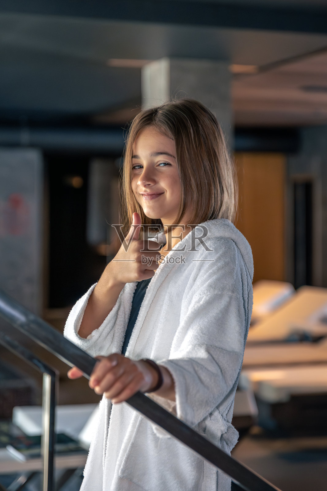 穿着白色浴袍、微笑的女孩在泳池旁的楼梯上竖起大拇指，像是刚游完泳。照片摄影图片