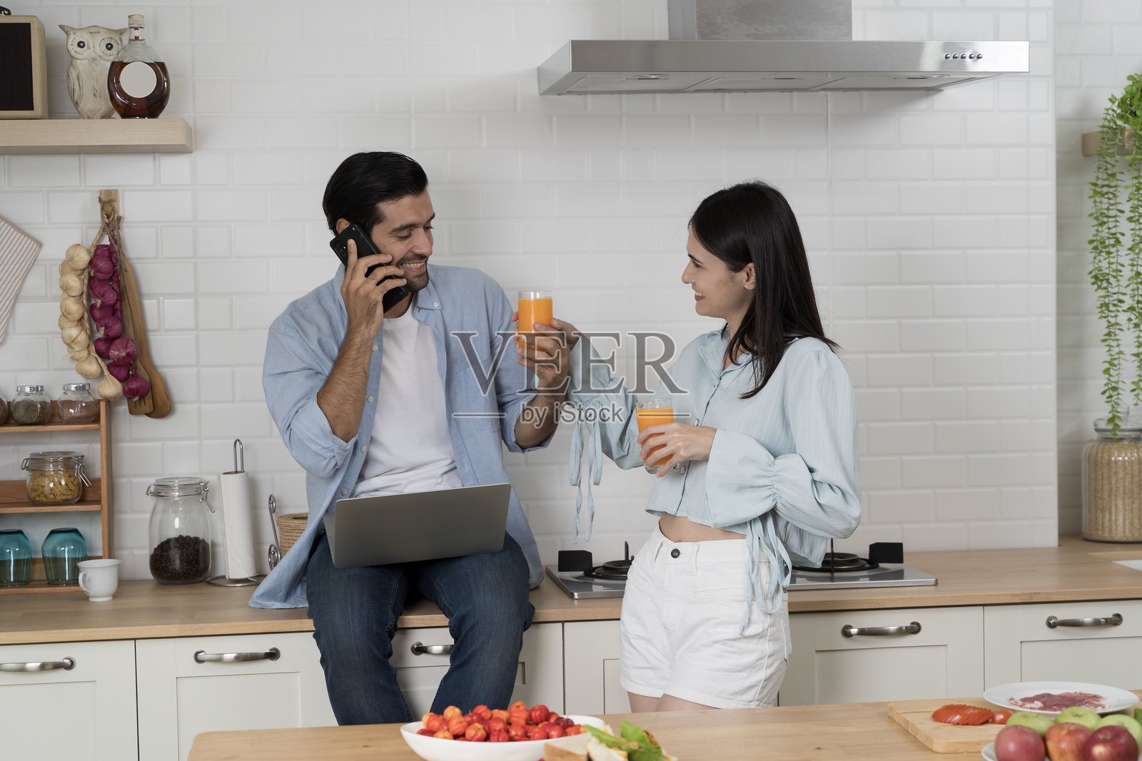 快乐的情侣在现代厨房里一起喝橙汁，男士一边打电话一边使用笔记本电脑，展现生活方式概念照片摄影图片