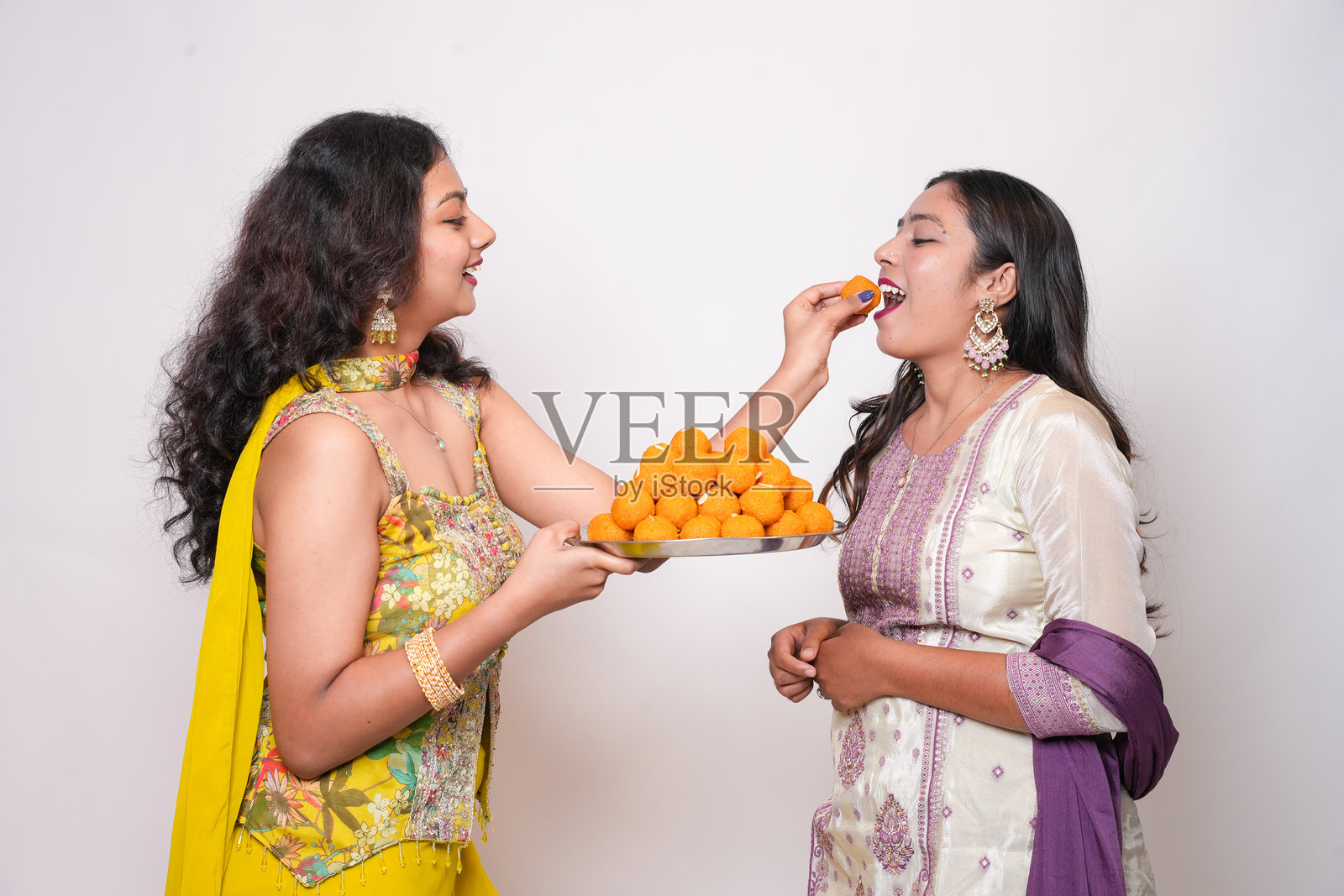 年轻的印度女性朋友或姐妹们在庆祝排灯节，享受吃拉杜甜点，背景为白色。照片摄影图片
