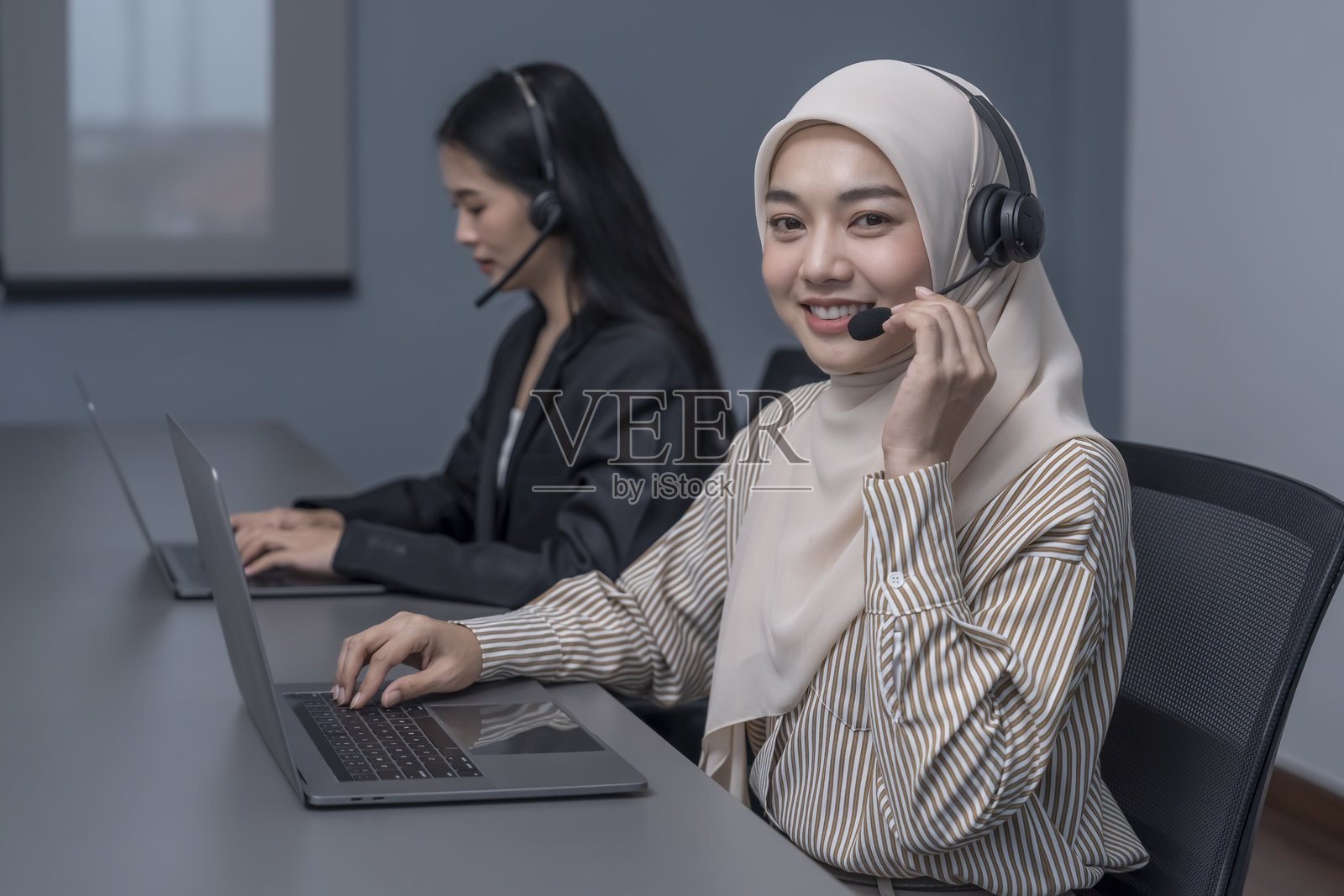 一位年轻微笑的女性戴着头巾，佩戴耳机，使用笔记本电脑，在呼叫中心工作，与客户交谈。照片摄影图片