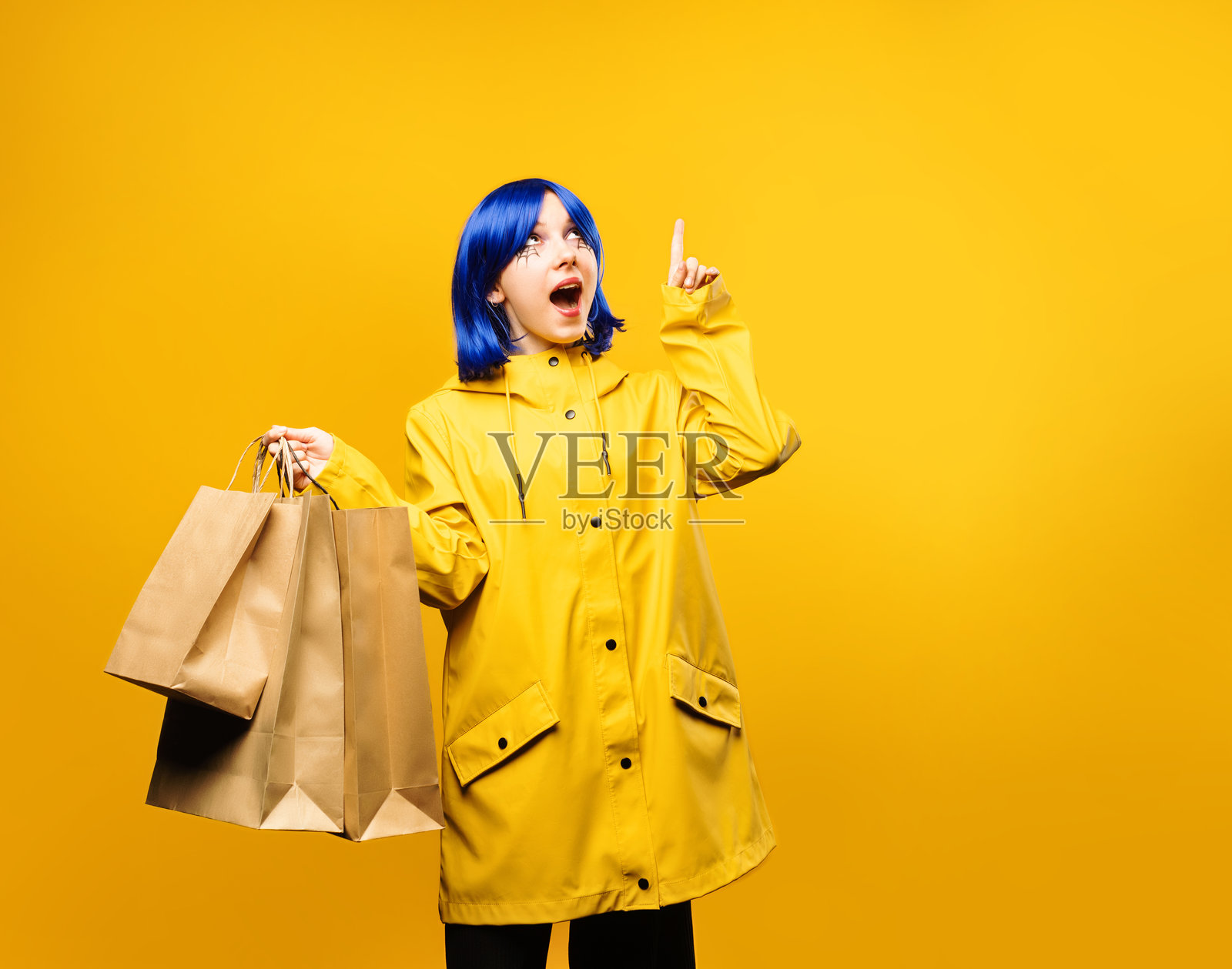 穿着黄色夹克的小女孩手持购物袋，指着黄色背景。万圣节促销或感恩节促销的概念，穿着《科拉琳》的服装。照片摄影图片