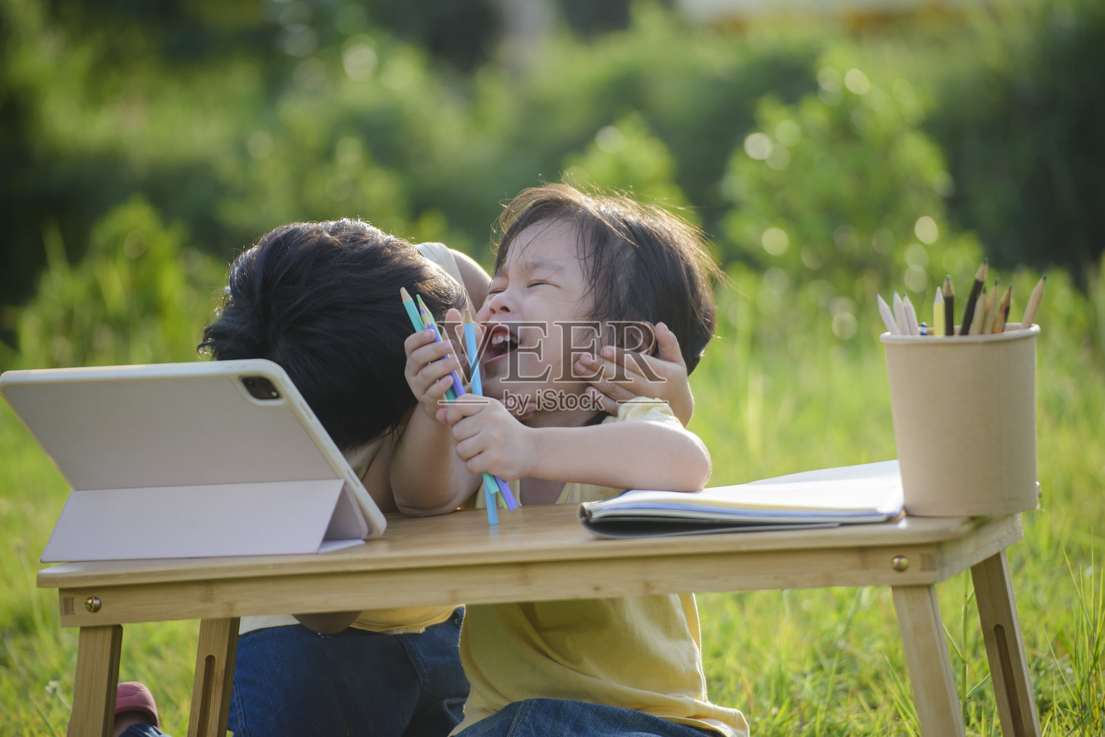 快乐的兄妹在户外一起学习。兄妹俩坐在木桌旁，手里拿着平板电脑和彩色铅笔，微笑着在阳光明媚的花园里一边学习一边玩耍。照片摄影图片