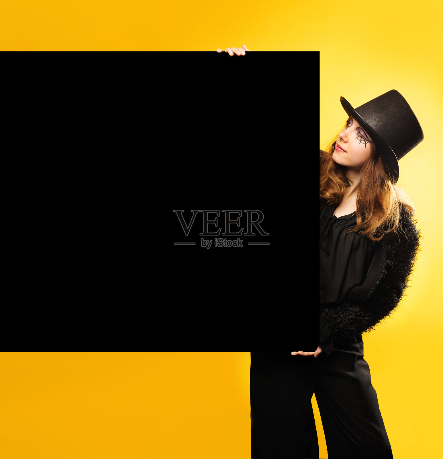 年轻女性穿着黑色万圣节服装，站在黄色背景前，手持黑色横幅或海报。留白空间。万圣节促销和派对概念。照片摄影图片
