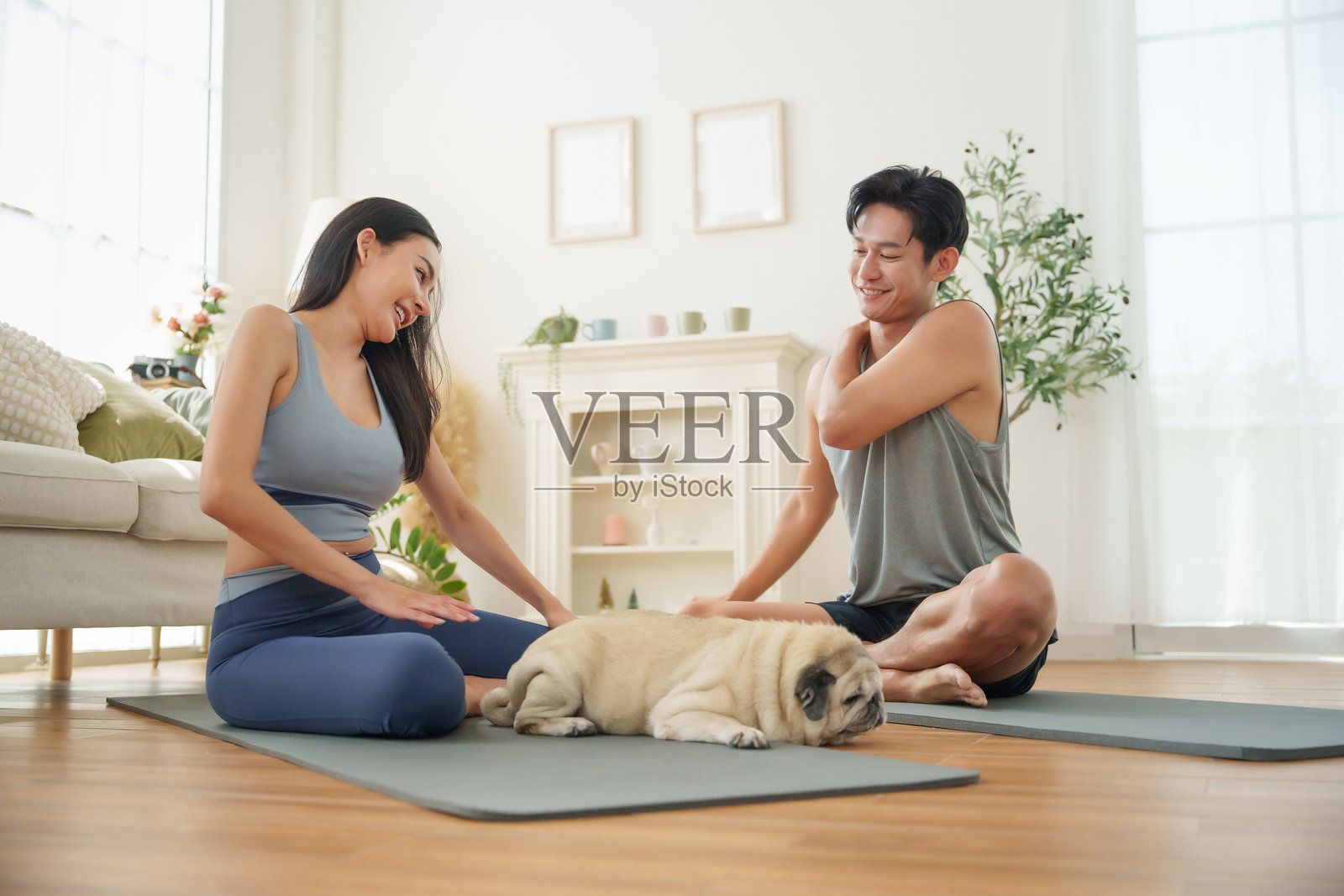 一对快乐的年轻亚洲夫妇在瑜伽垫上放松，他们的巴哥犬在旁边悠闲地躺着，分享着愉快的微笑，刚刚一起完成了一次有趣的家庭锻炼。照片摄影图片