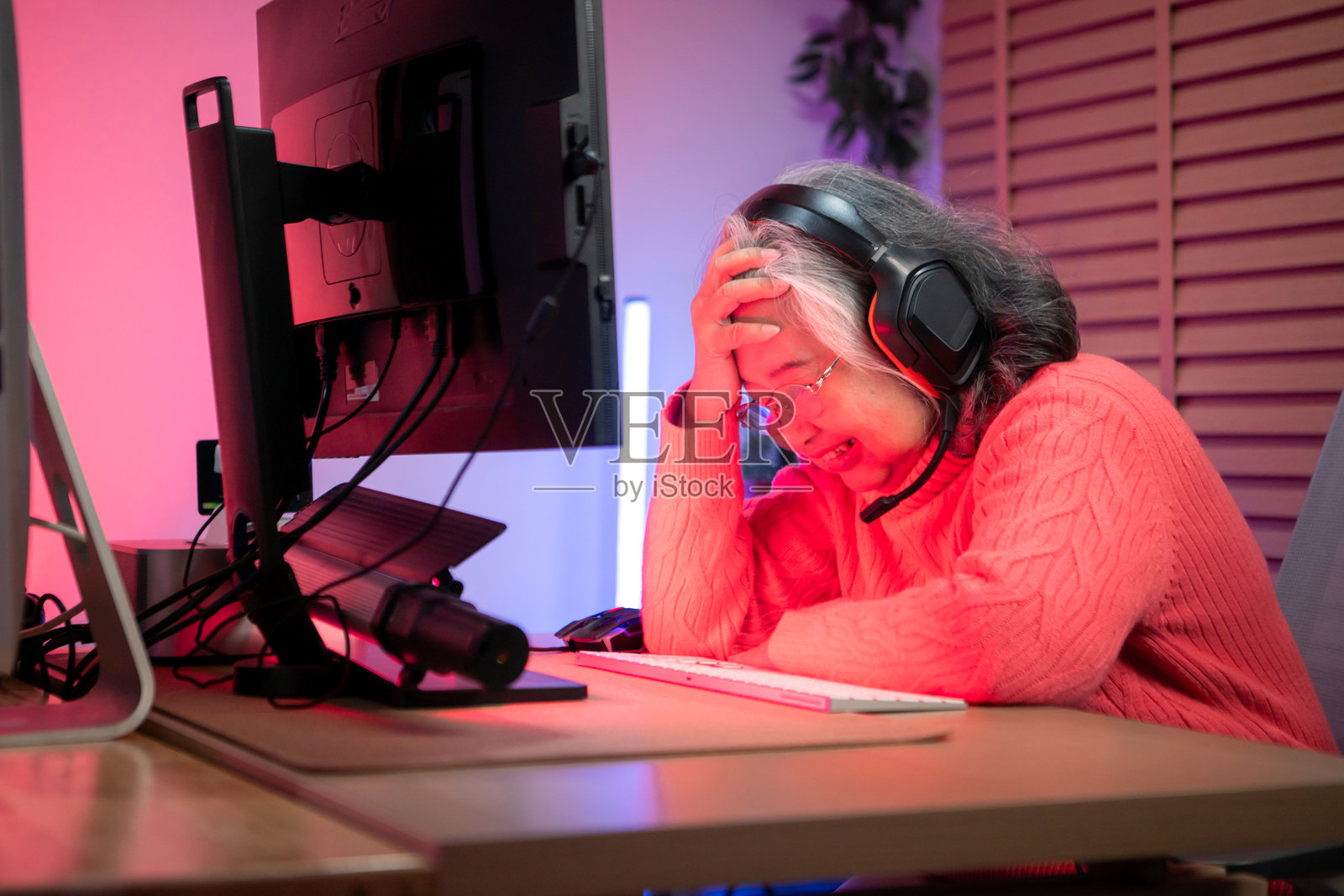 戴着耳机的老年女性玩家在色彩缤纷的游戏设置中使用台式电脑游戏照片摄影图片