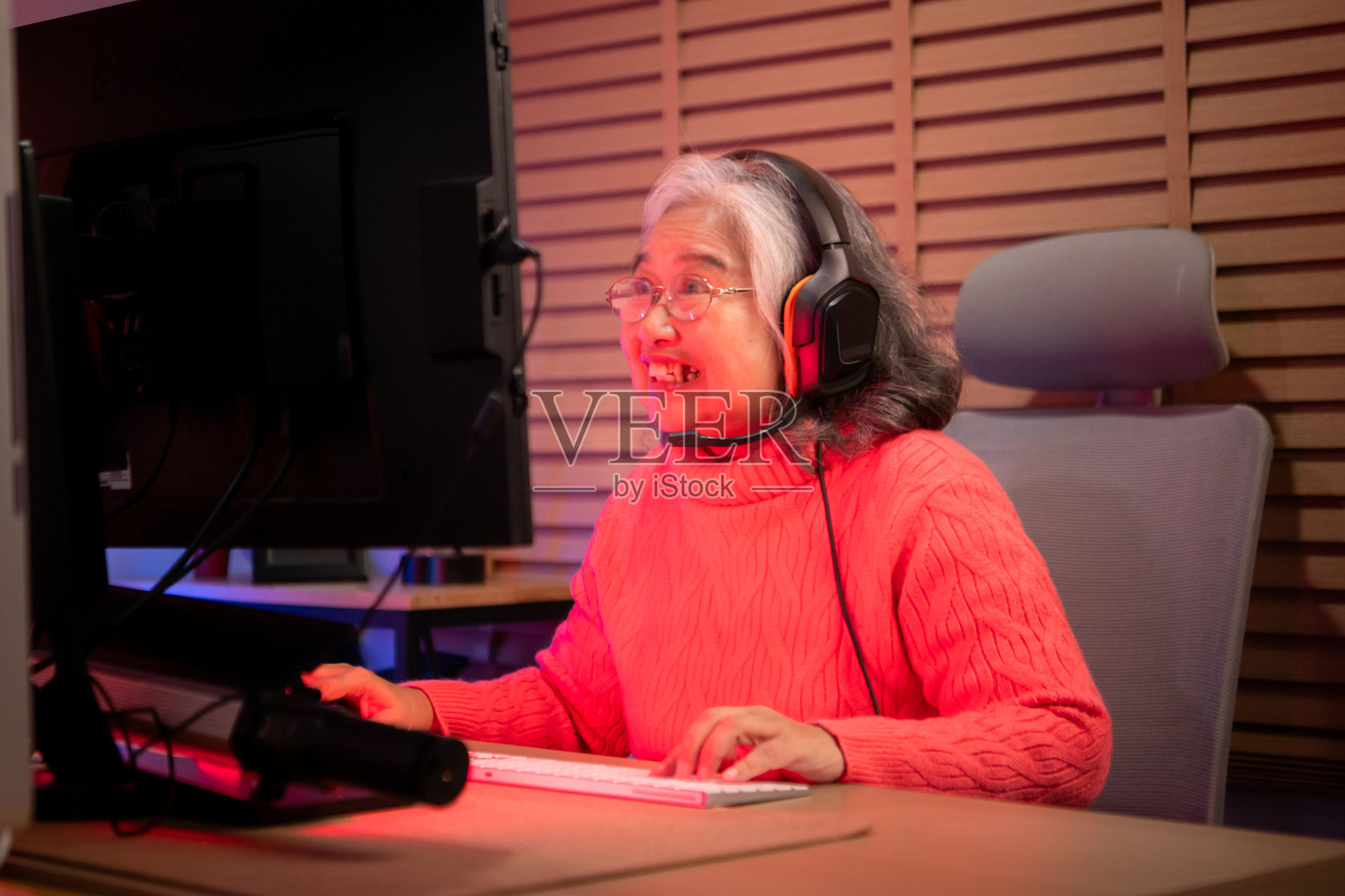 一位年长女性玩家戴着耳机，在色彩缤纷的游戏设置中使用台式电脑游戏。照片摄影图片