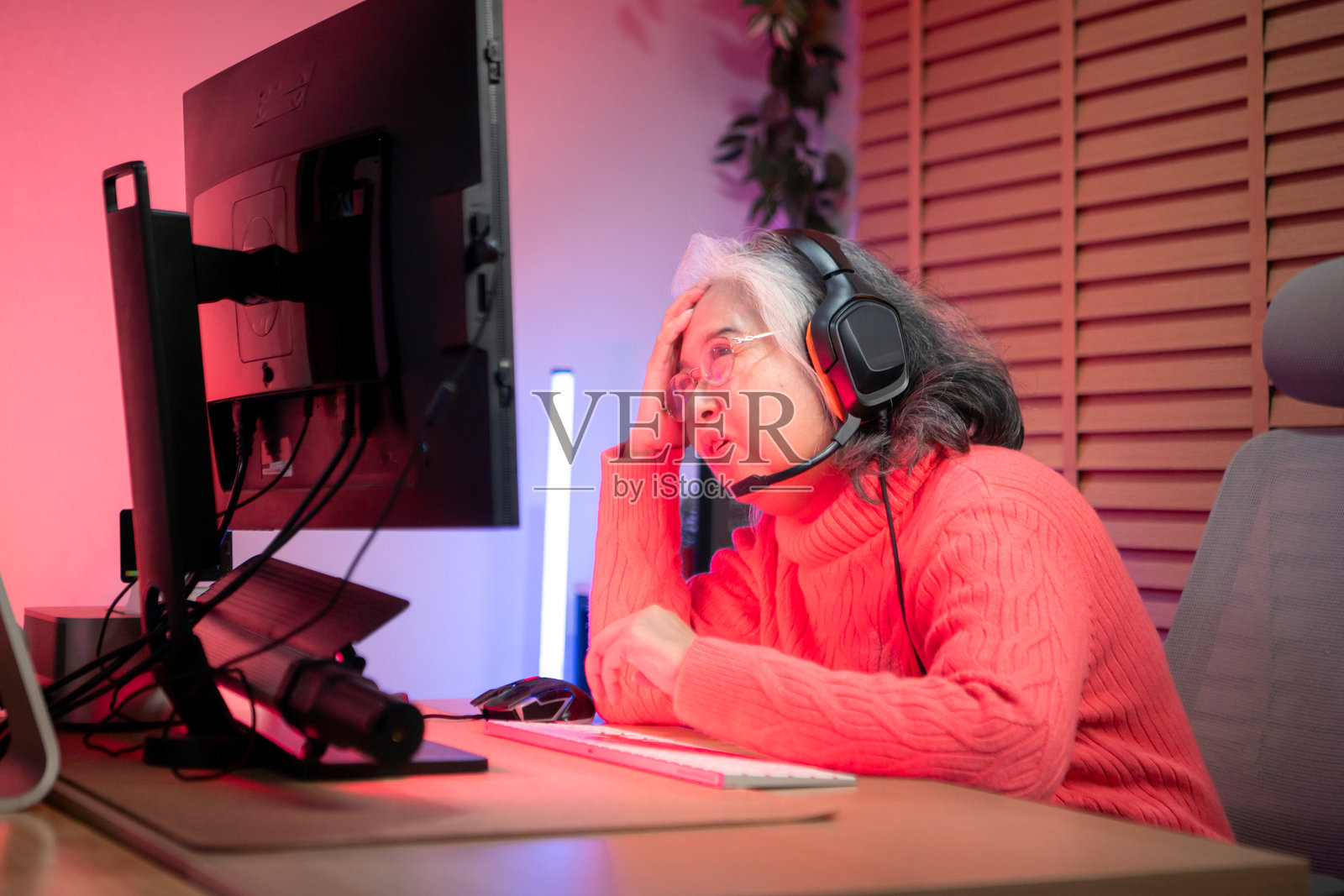 戴着耳机的老年女性玩家在色彩缤纷的游戏设置中使用台式电脑游戏照片摄影图片