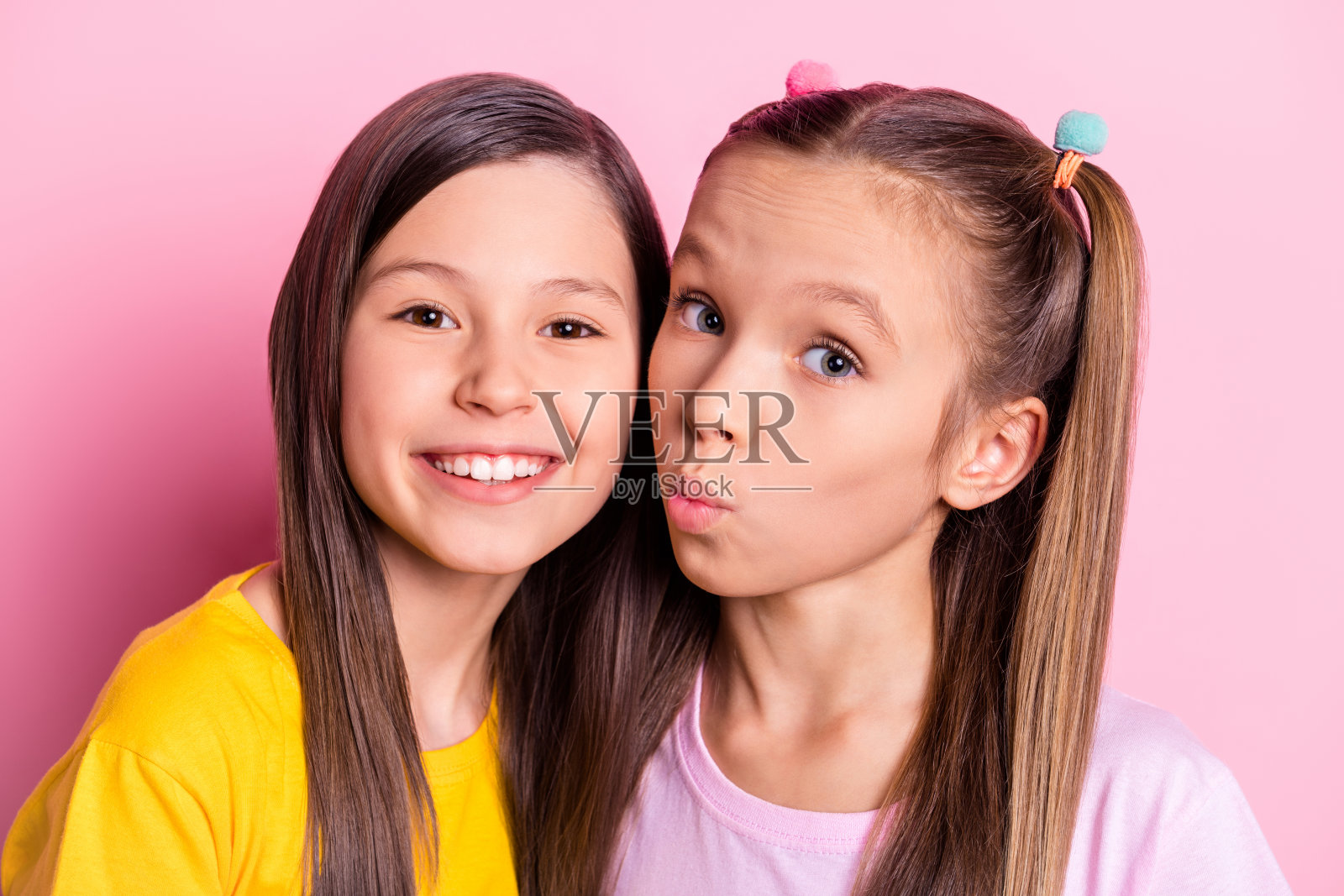 乐观的棕发和金发女孩亲吻，穿着黄色和粉色T恤，背景为柔和的粉色。照片摄影图片