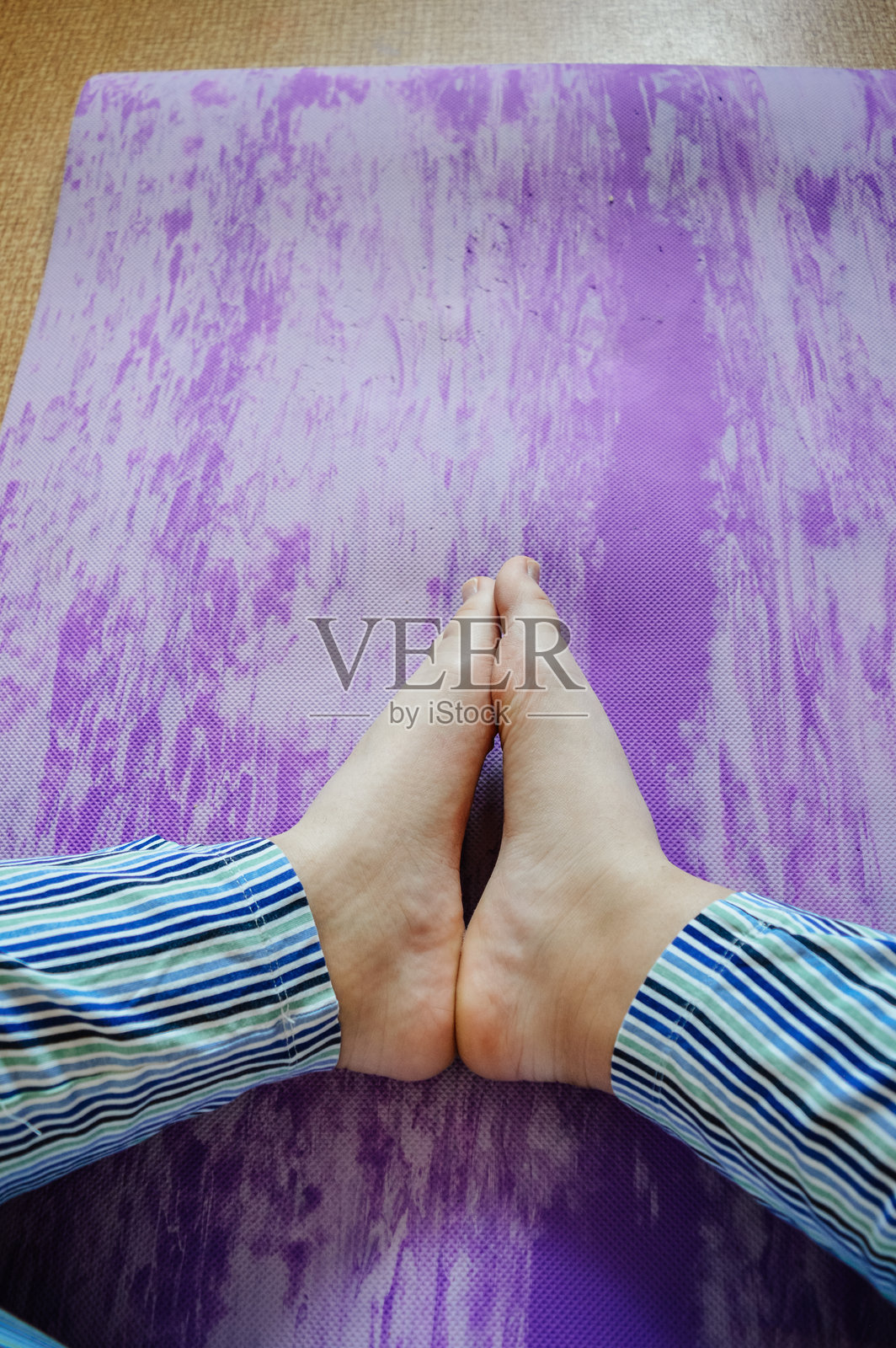 一个人盘腿坐在紫色瑜伽垫上，双脚并拢，穿着条纹裤。冥想、放松、瑜伽练习和健康生活方式的概念。照片摄影图片