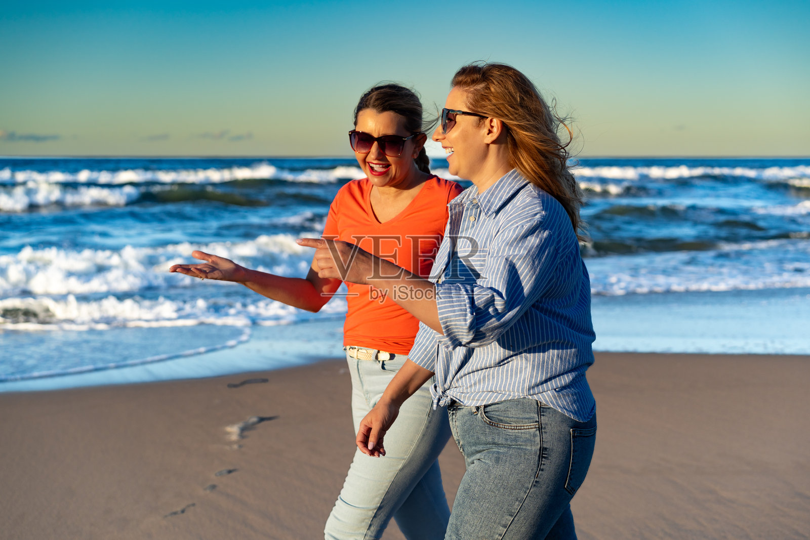 夏季假期。两位美丽成熟的女性在阳光明媚、微风拂面的沙滩上散步。照片摄影图片