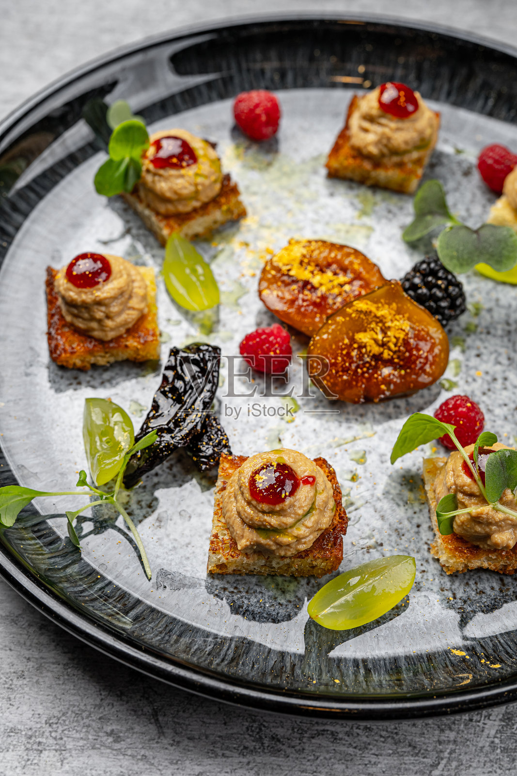 优雅的美食开胃菜盘，配有各种顶层为肉酱和浆果酱的迷你点心、焦糖水果、微型绿叶、鲜覆盆子、黑莓和葡萄，摆放在黑色陶瓷盘上。照片摄影图片