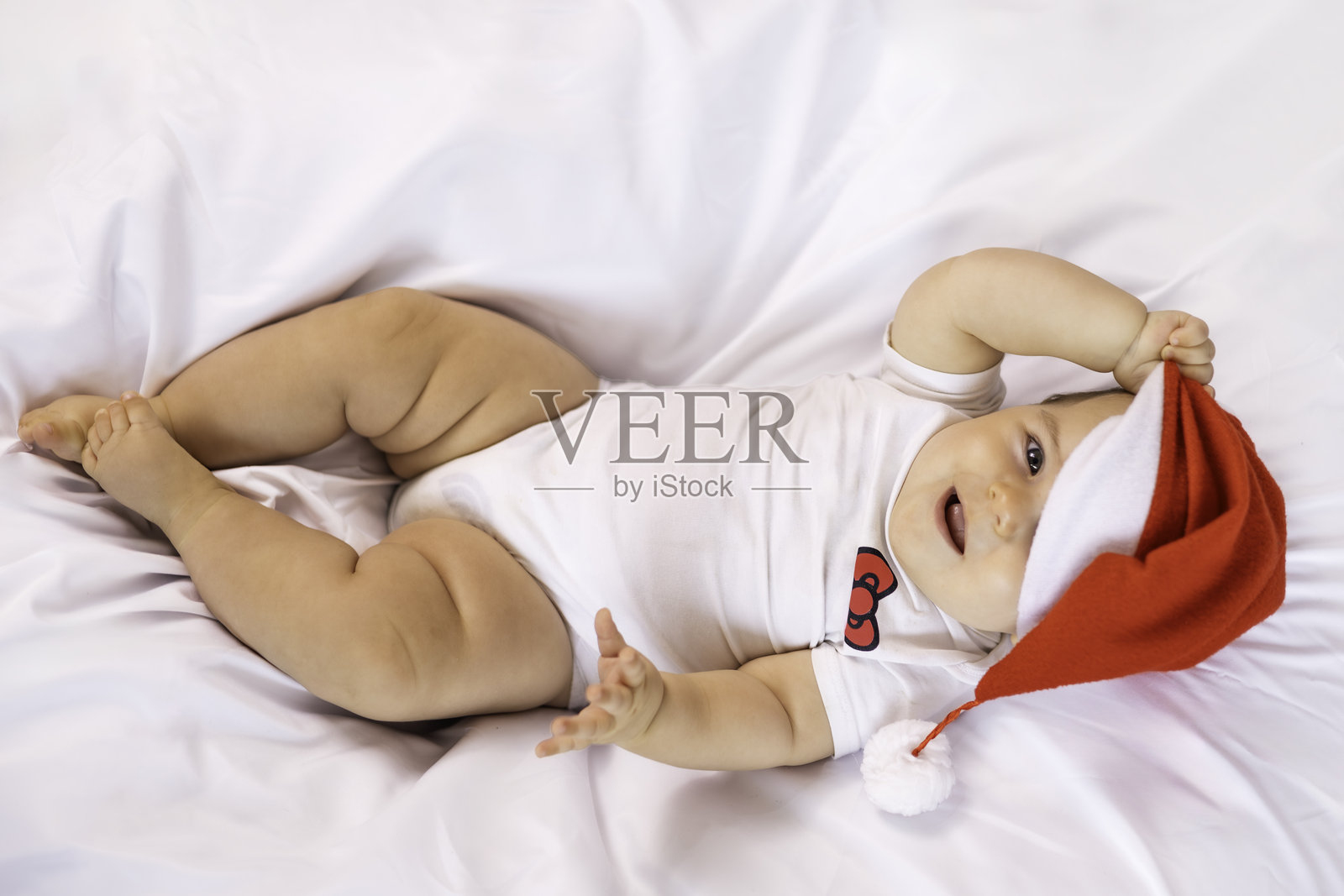 小圣诞老人矮人。一个戴着圣诞帽的小宝宝，躺在白色床单上，穿着白色连体衣，衣服上系着红色丝带。照片摄影图片