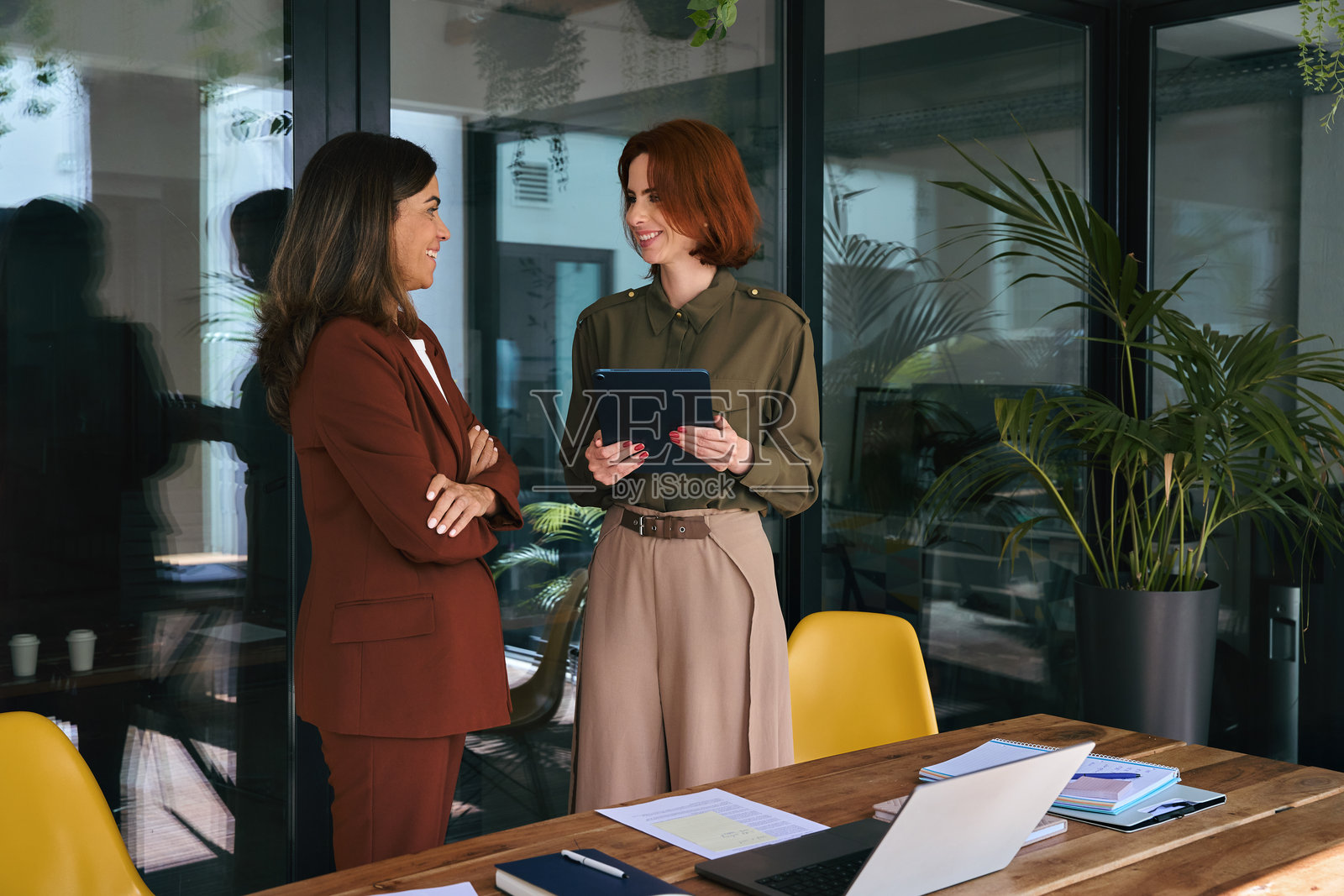 两位忙碌快乐的女商人在办公室里站着用平板电脑交谈。照片摄影图片