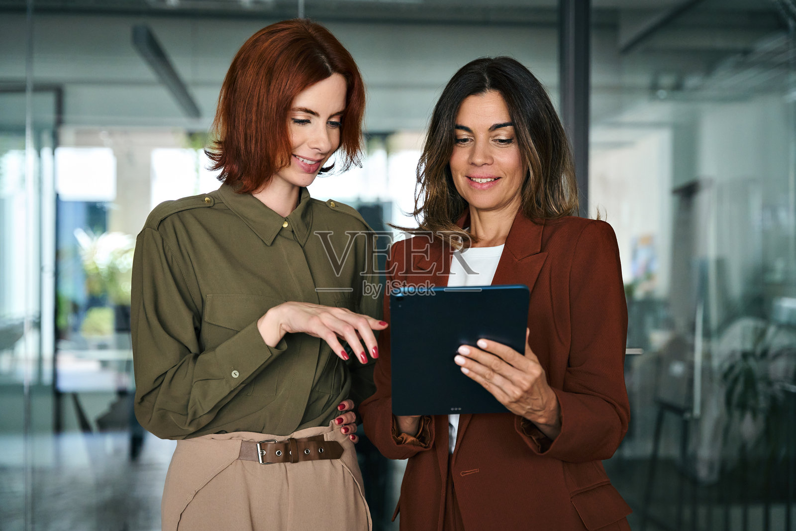 两位忙碌快乐的女商人在办公室里站着交谈，使用平板电脑。照片摄影图片