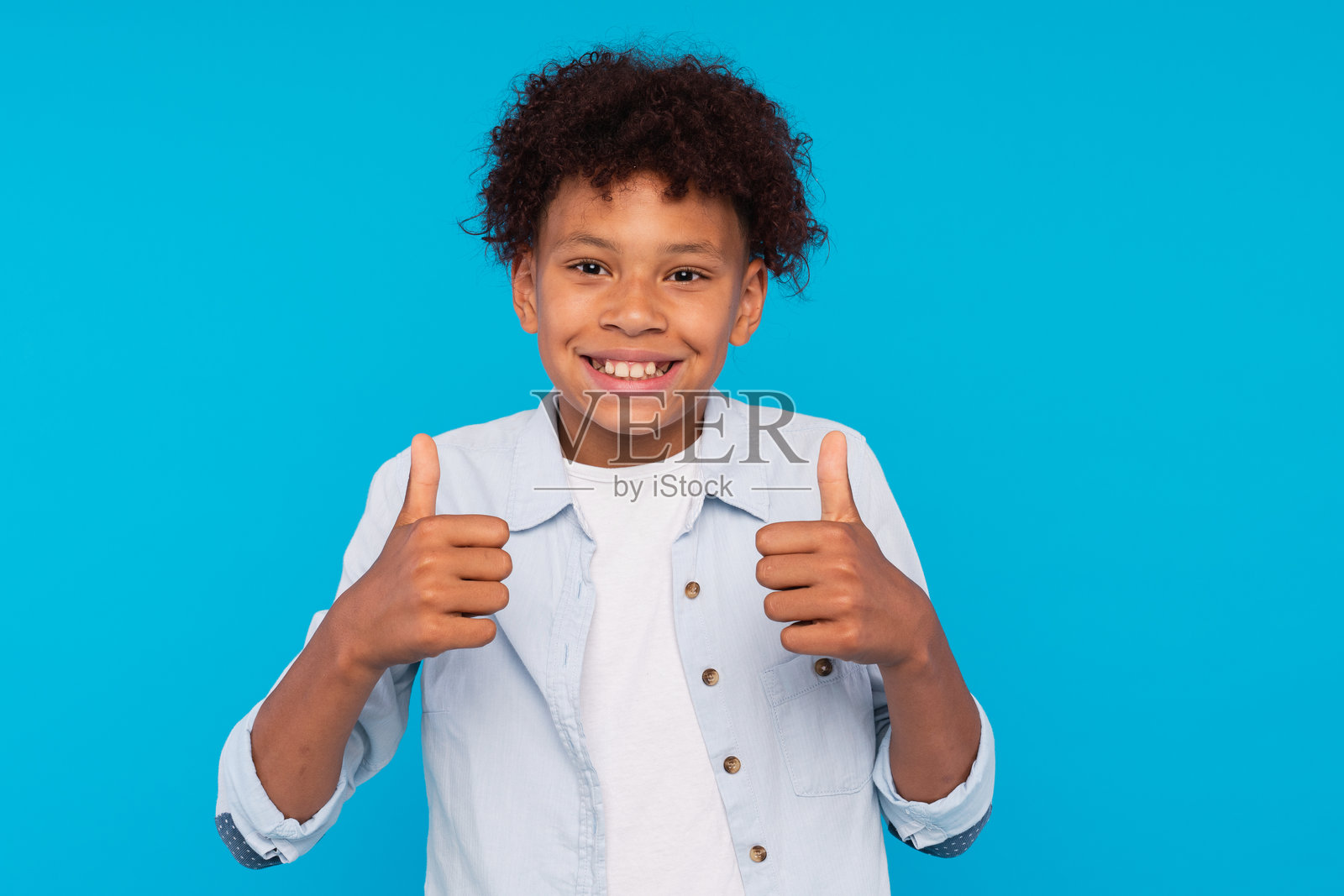 快乐的小男孩肖像，卷发，穿着白色T恤，微笑着看向镜头，做出竖起大拇指的手势，表示赞同和酷炫的认可。室内摄影，背景为蓝色。照片摄影图片