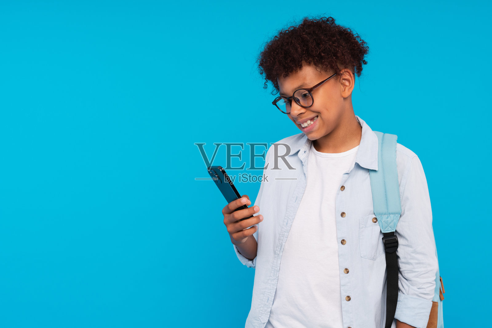 微笑的非裔美国小学生男孩，背着书包，手持智能手机，正在使用互联网移动应用程序发送短信，目不转睛地看着屏幕，背景留白。照片摄影图片