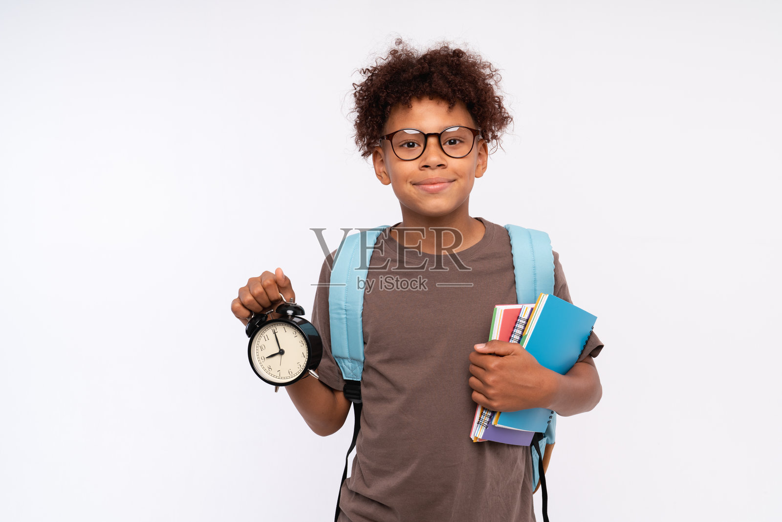 微笑的非裔美国男孩，初中生，背着书包，手握圆形时钟，背景为白色。教育学术，九月一日开学的概念。照片摄影图片