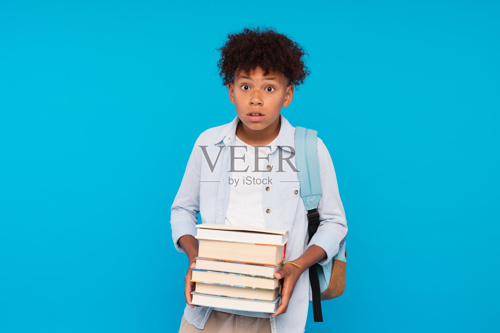 惊讶的小黑人男孩，背着书包，手里拿着教育书籍，背景为蓝色。九月一日开学的教育学术概念。照片摄影图片