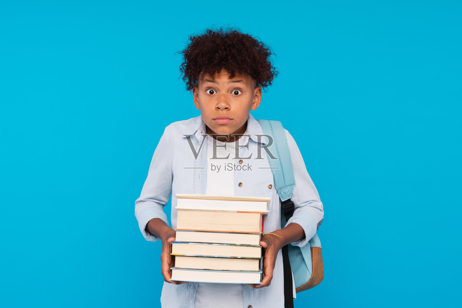 惊讶的非裔美国男孩，背着书包，手里拿着沉重的书籍，孤立在蓝色背景下。教育回到学校的概念。照片摄影图片