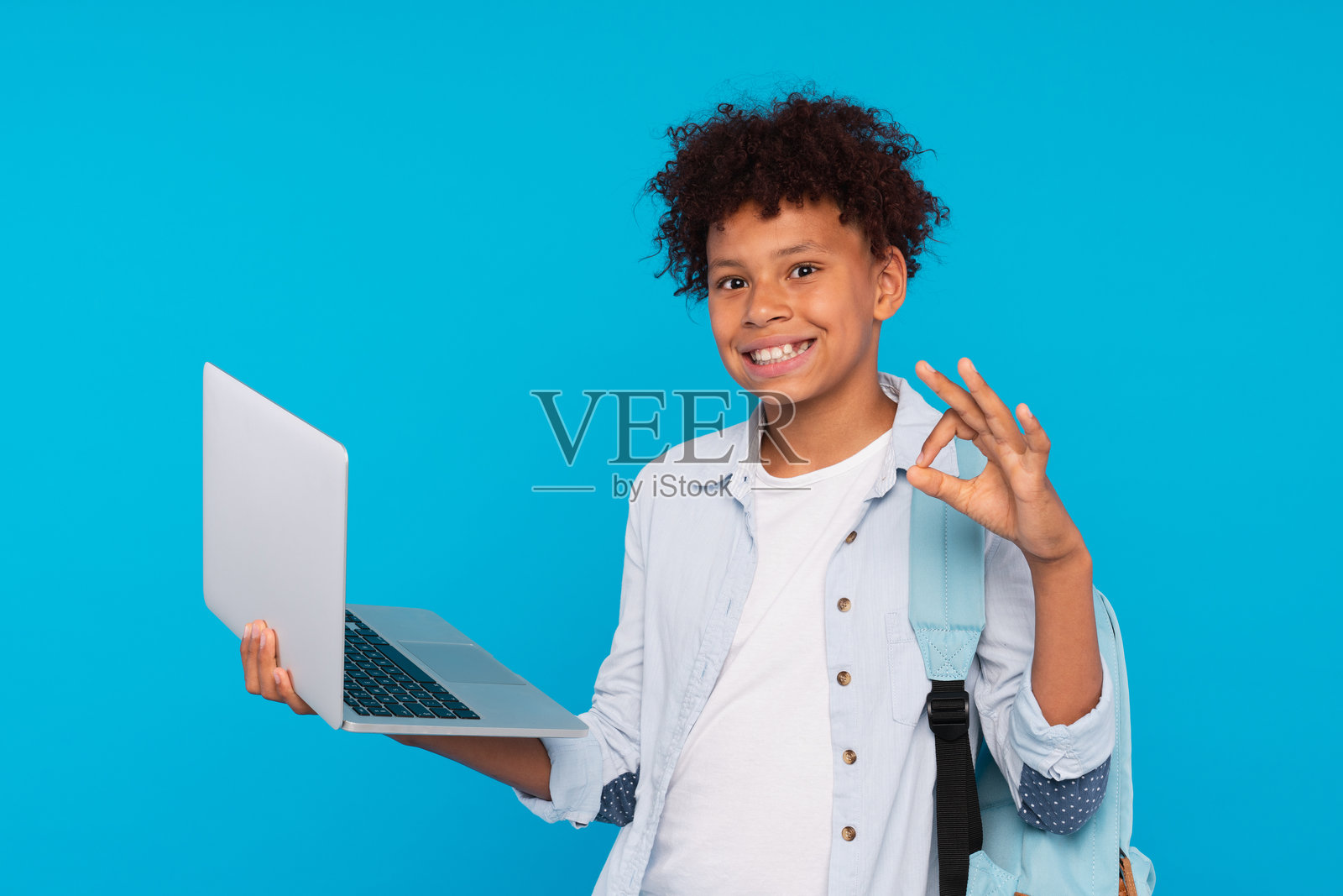 非裔美国小学生男孩展示“好”手势，手持笔记本电脑，正在上网冲浪，远程学习，背景为蓝色。照片摄影图片