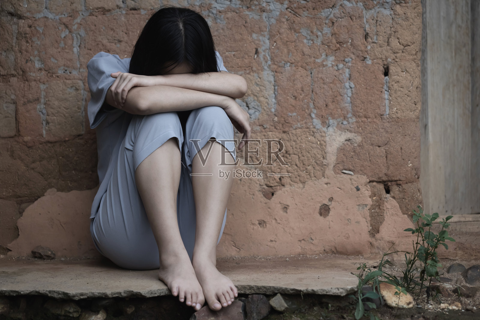 一个贫穷、沮丧的年轻女孩坐在一面古老的砖墙旁，低着头哭泣。照片摄影图片