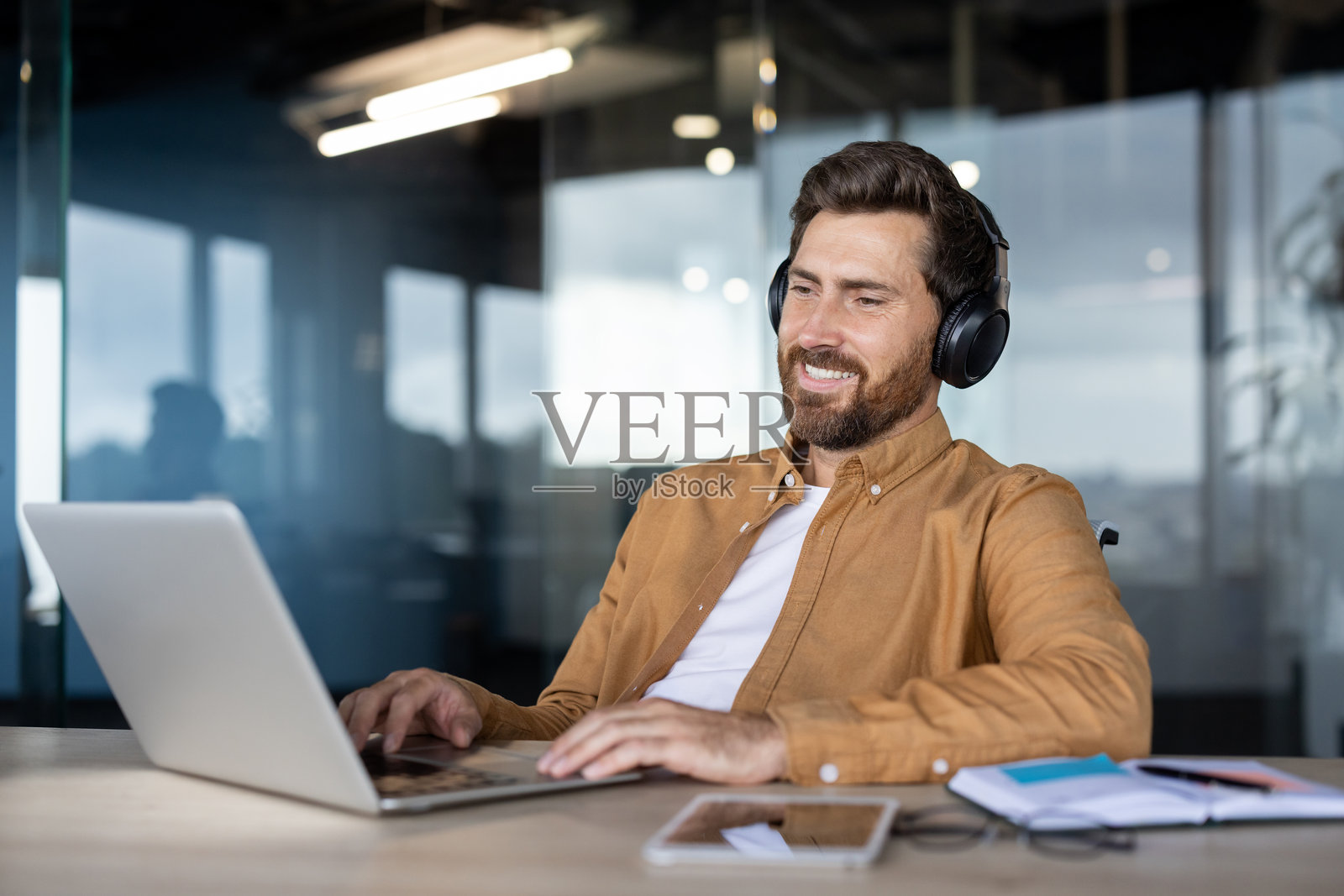 一个快乐的男人戴着耳机在现代办公室环境中使用笔记本电脑。照片摄影图片