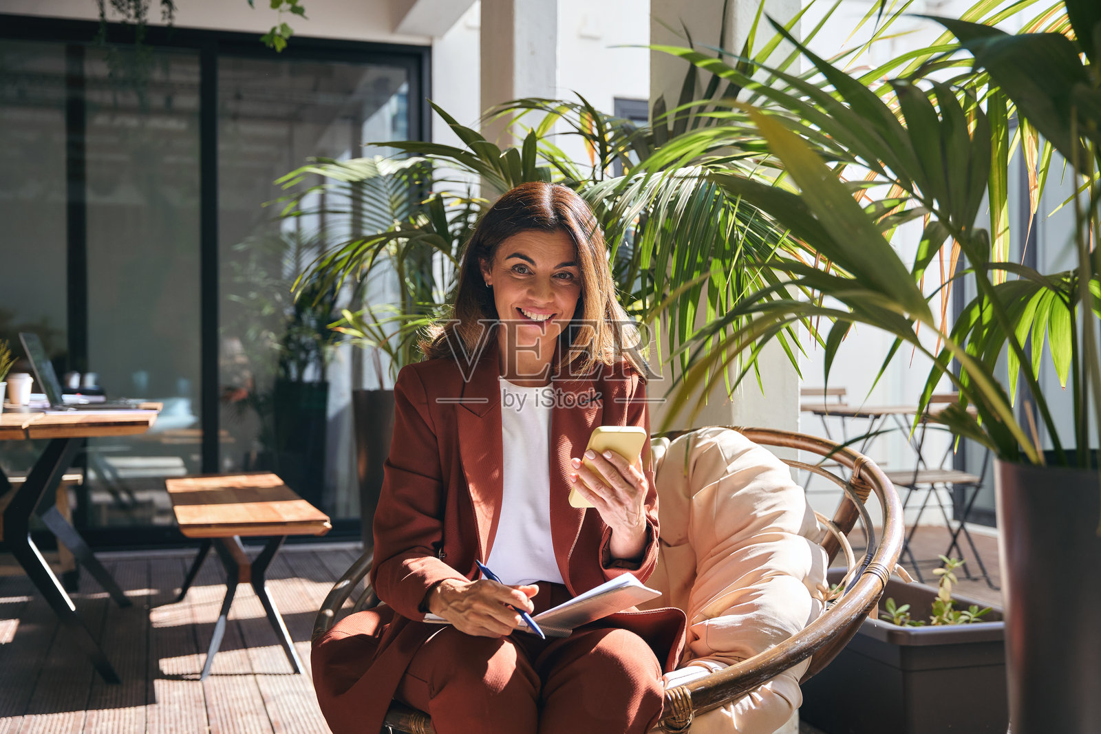 快乐的成熟的女性商务人士坐在椅子上，阳光明媚的绿色办公室里使用手机。照片摄影图片