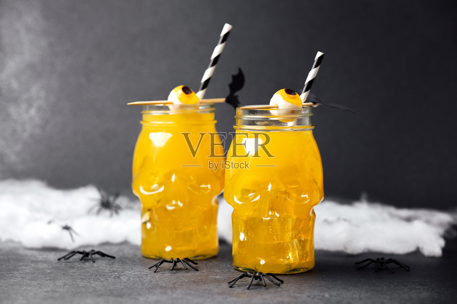 两个装满黄色饮料、冰块、吸管和糖果眼球的骷髅杯，放在黑色表面上，周围有蛛网和蜘蛛。照片摄影图片