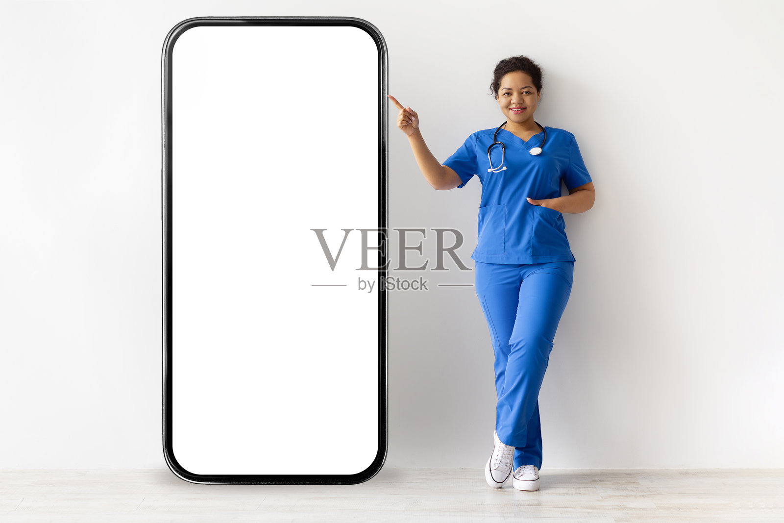 穿着护士服的黑人女性护士，手持听诊器，站在大型智能手机模型旁边照片摄影图片