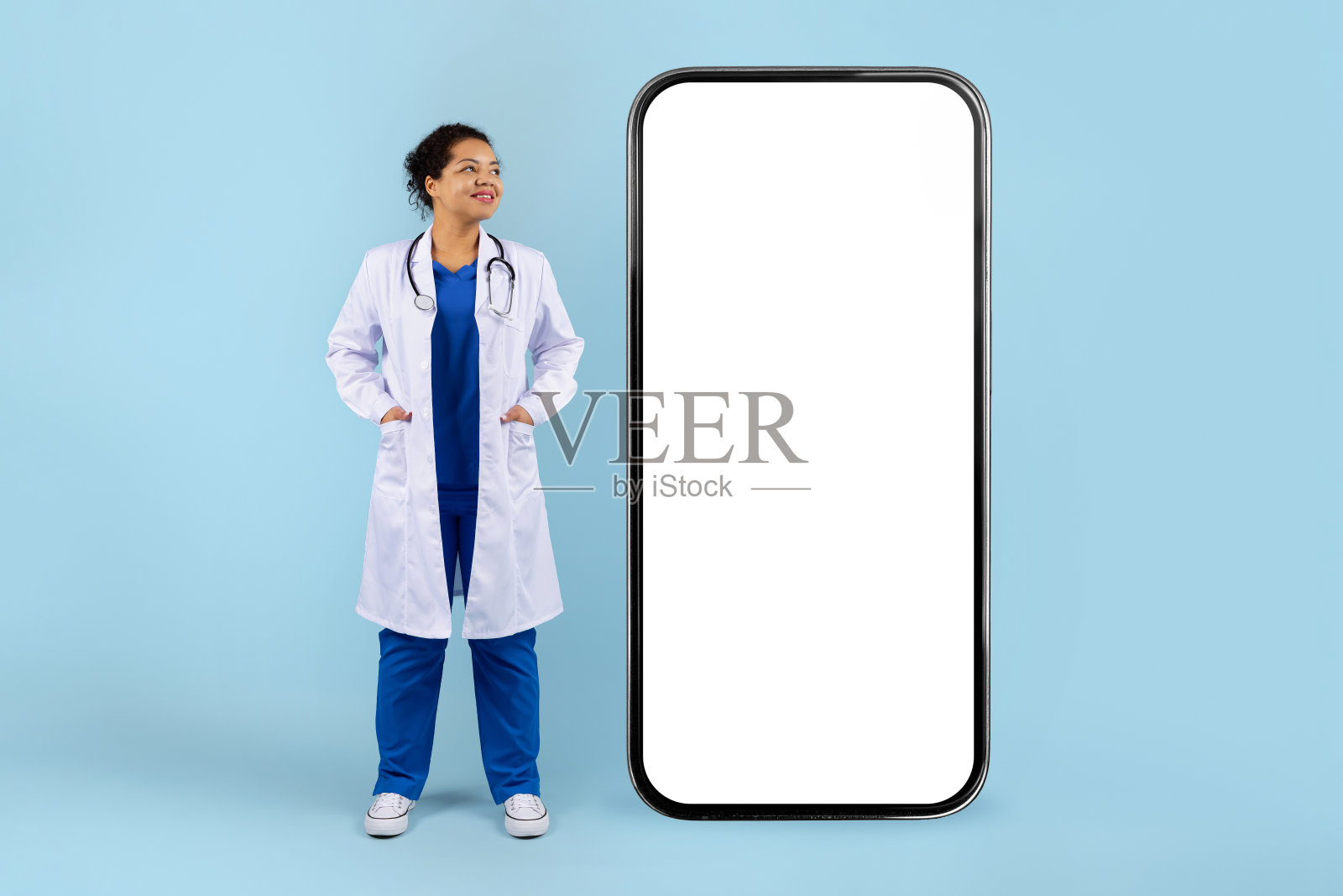 穿着实验室外套的黑人女性医生站在大型智能手机模型旁边照片摄影图片