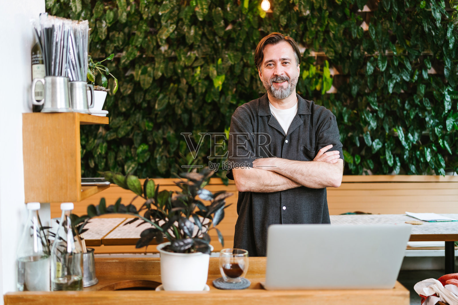 小企业主站在咖啡馆柜台前，双臂交叉，背景是绿植墙和咖啡杯，现代环保的工作空间氛围。照片摄影图片