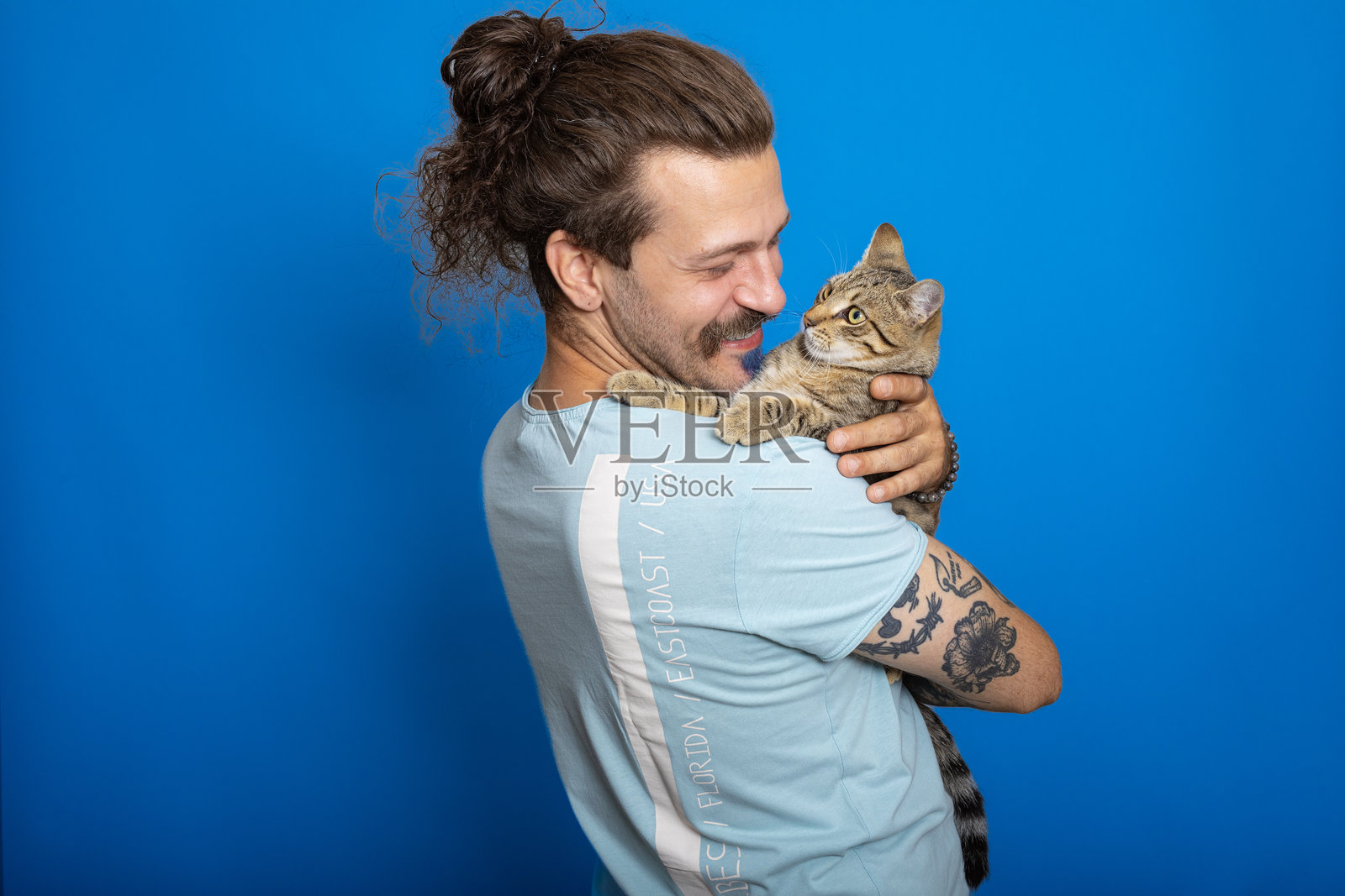 快乐的男人穿着休闲T恤，抱着他的猫咪，背景为蓝色照片摄影图片