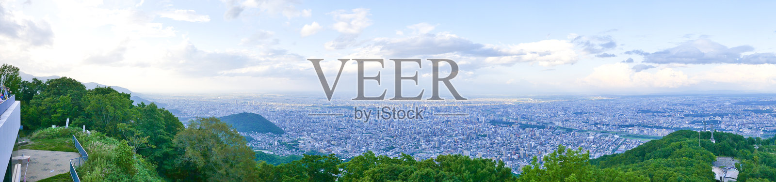 从藻岩山的全景视角可以看到札幌市在风景中延展，四周被郁郁葱葱的山丘环绕，天空布满云彩。照片摄影图片