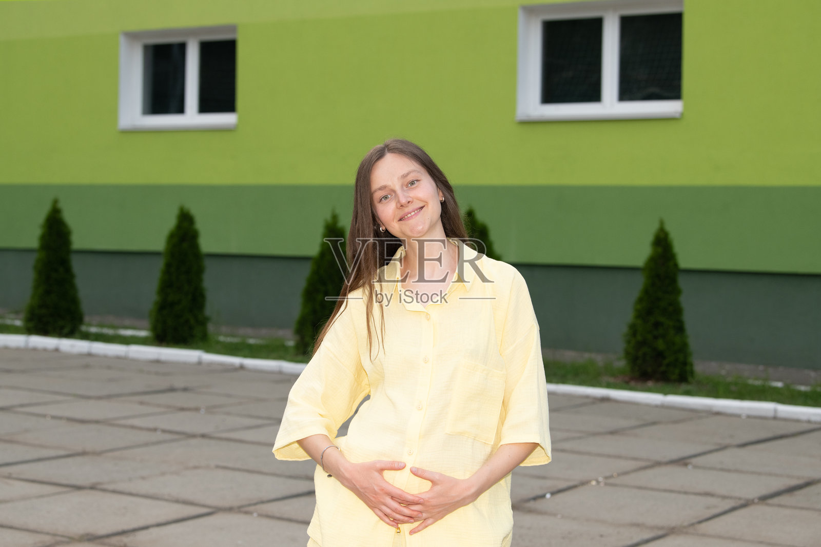 一位年轻女性在绿色建筑前的户外站着。她温暖地微笑着，轻轻地抚摸着自己的肚子，享受着傍晚阳光下的宁静时刻。照片摄影图片