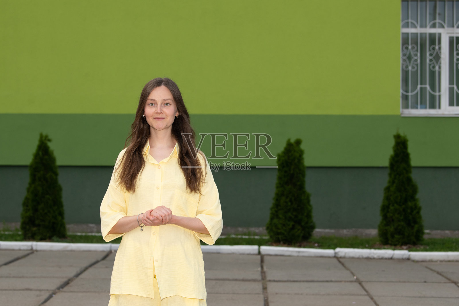 一位穿着黄色服装的年轻女性在户外一面生机勃勃的绿色墙前温暖地微笑。这个场景传达出积极和清新的感觉。照片摄影图片