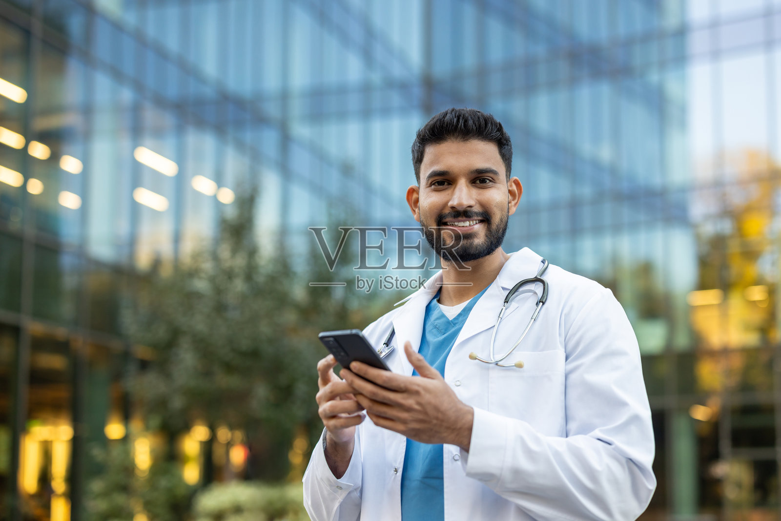 一位微笑的医生在现代建筑旁拿着手机看向镜头。照片摄影图片
