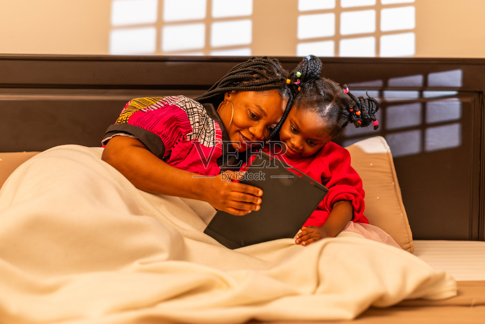 非洲母亲和孩子在夜晚的被窝里亲密互动，一起在平板电脑上观看视频。照片摄影图片