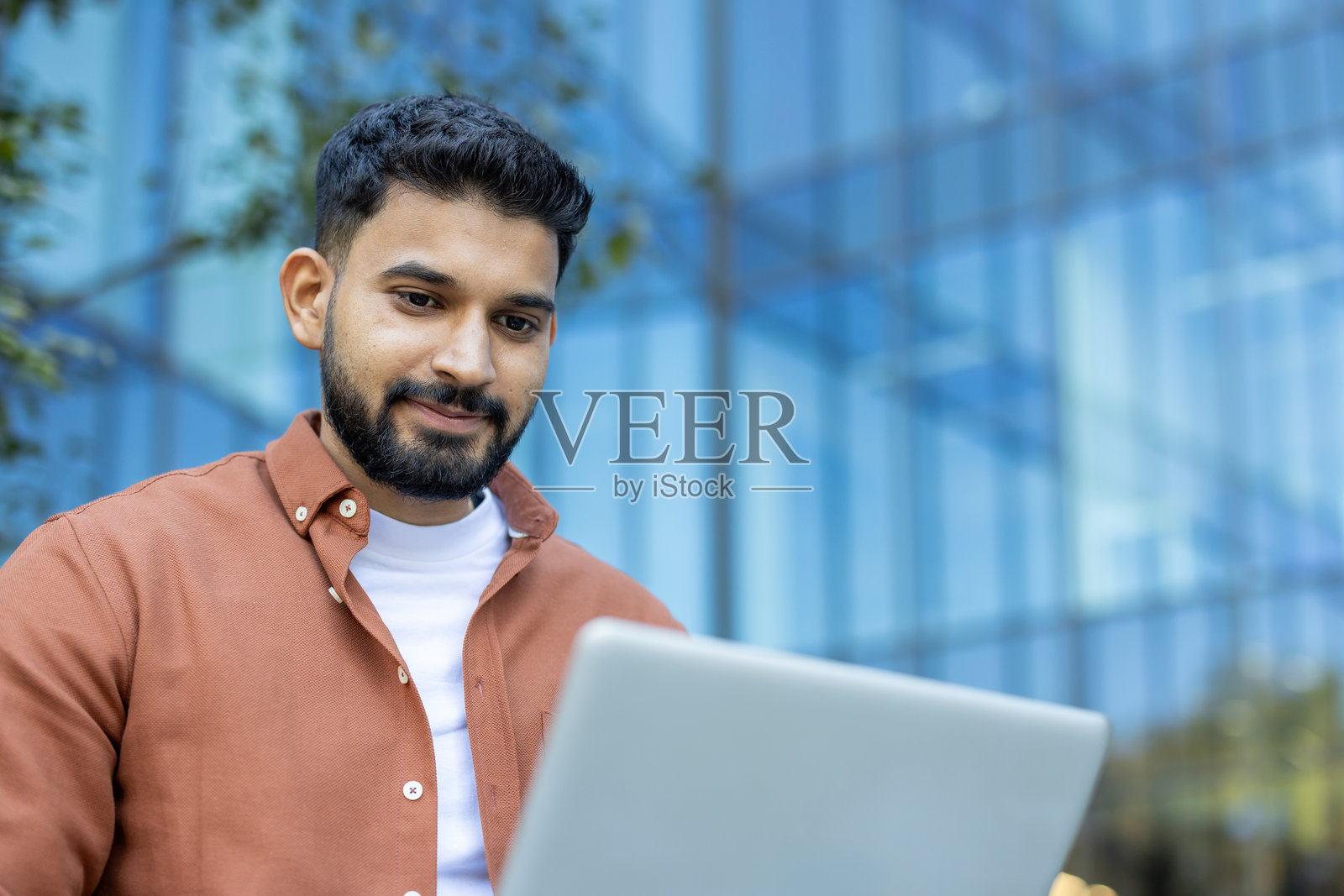 一位年轻的印度男子在户外阳光下微笑着使用笔记本电脑。照片摄影图片