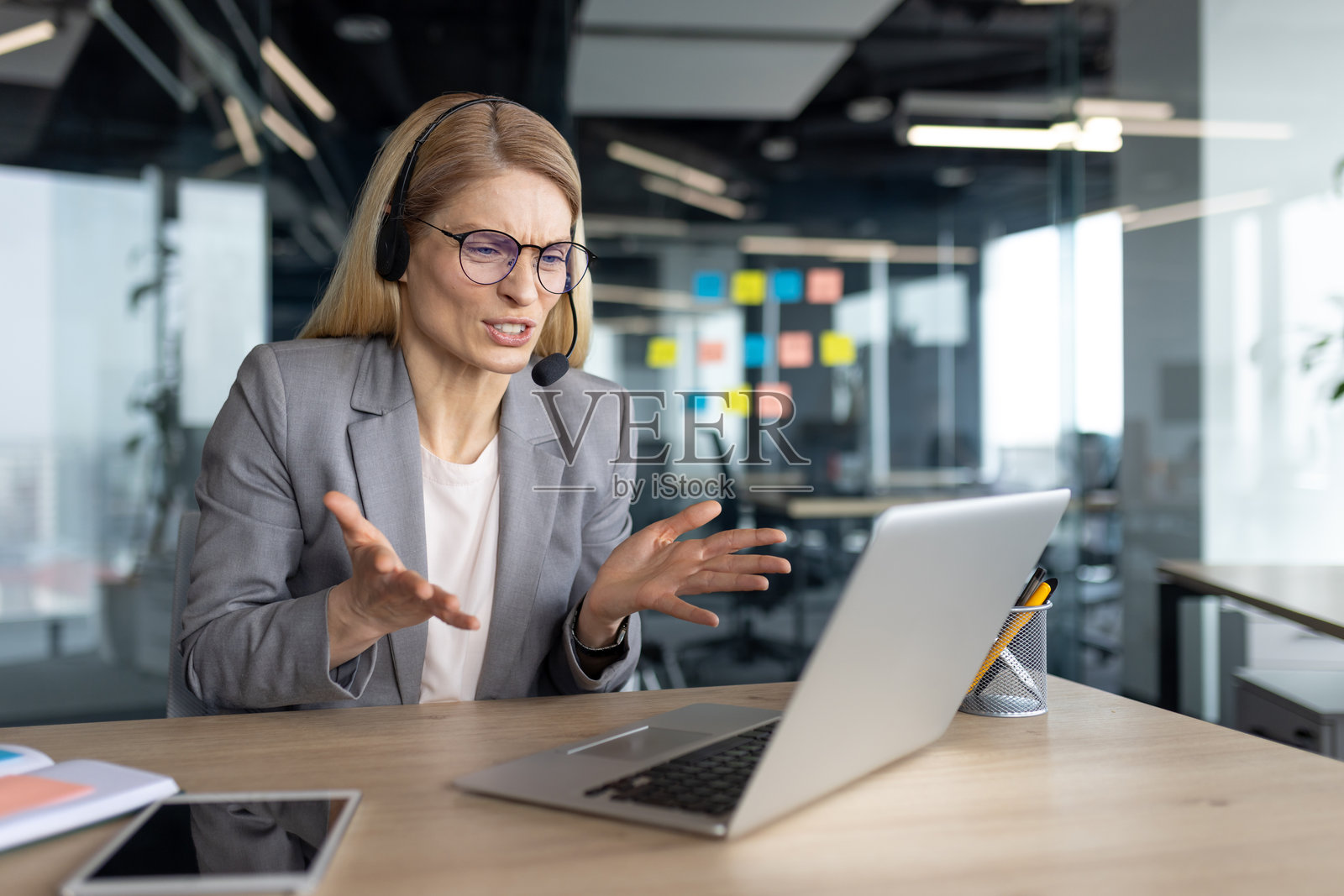 紧张且愤怒的女性坐在办公室里，使用耳机电话和笔记本电脑进行视频通话。身穿商务套装的女商人在在线商务会议中。照片摄影图片