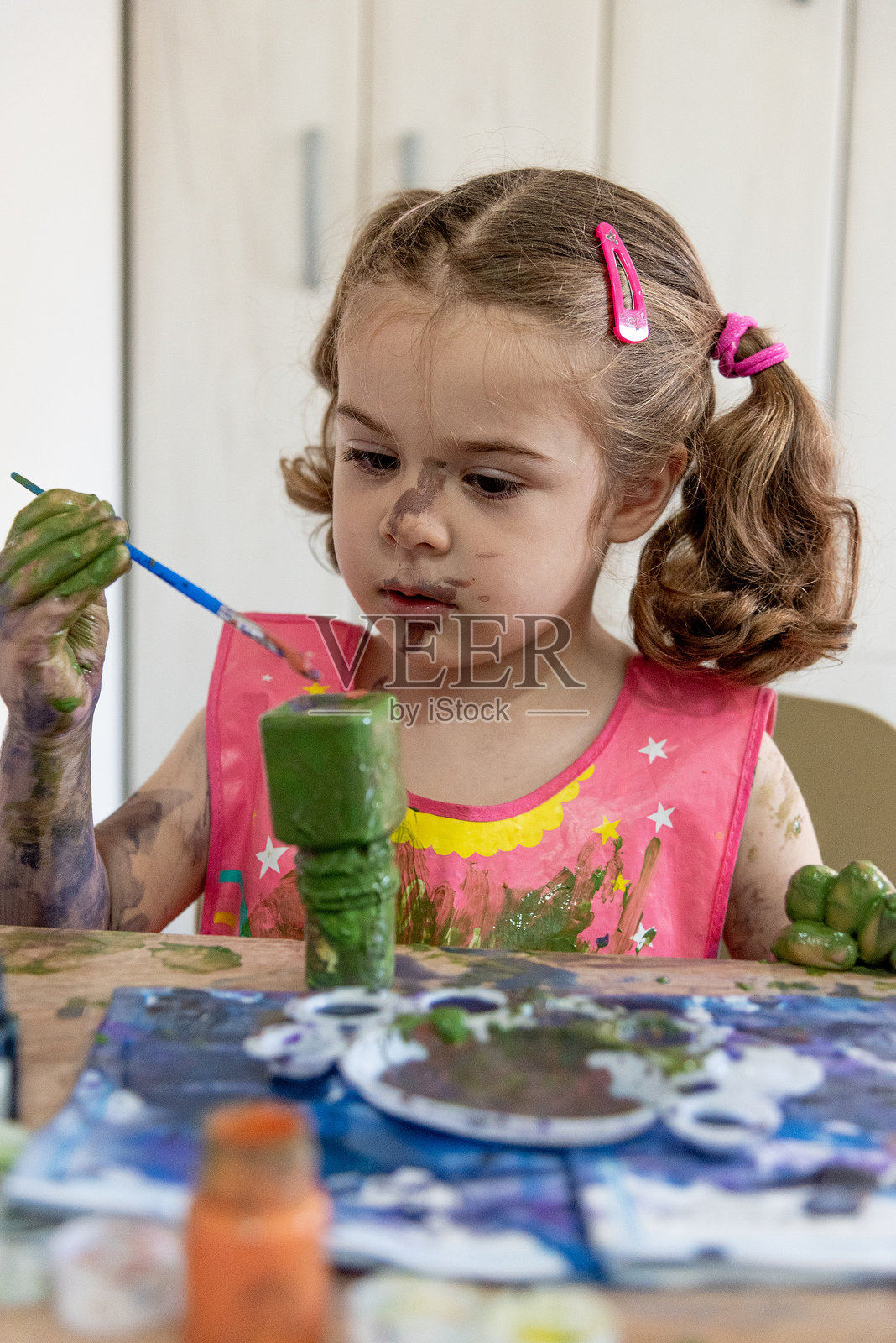 小女孩穿着围裙，用彩色颜料在儿童涂色书上画画，孩子的手上沾满了水粉颜料。照片摄影图片