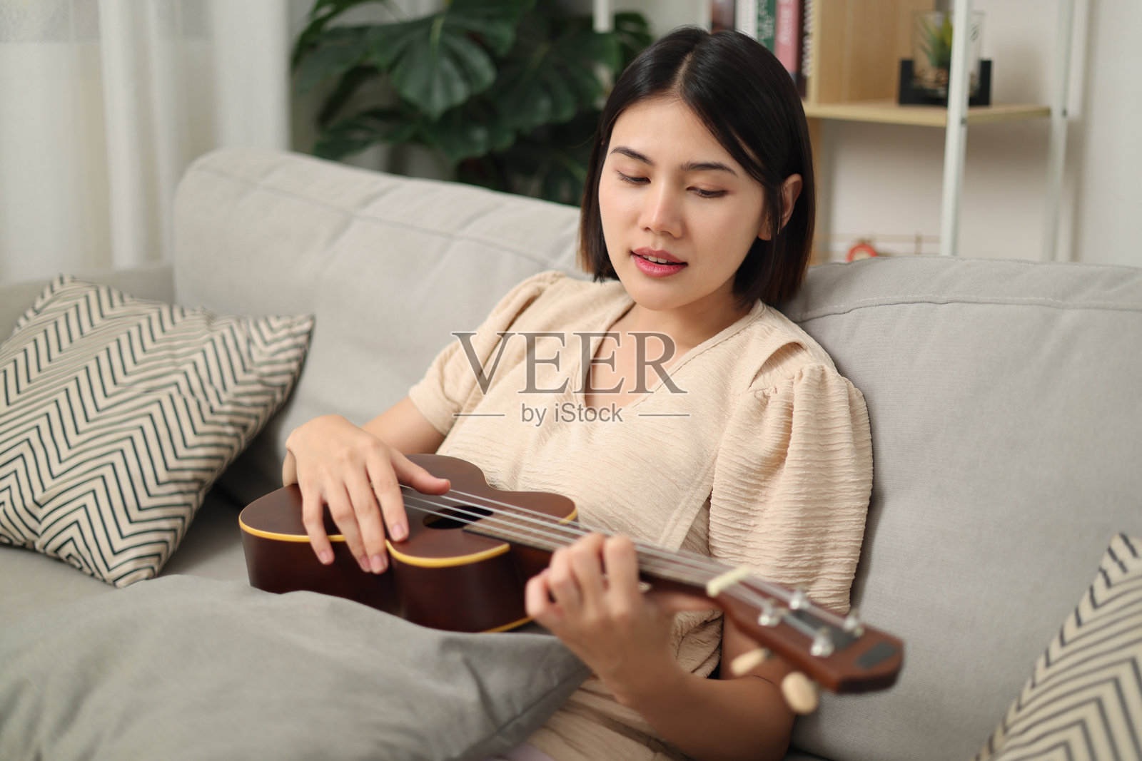 年轻的亚洲女性在家里的沙发上享受弹尤克里里。照片摄影图片