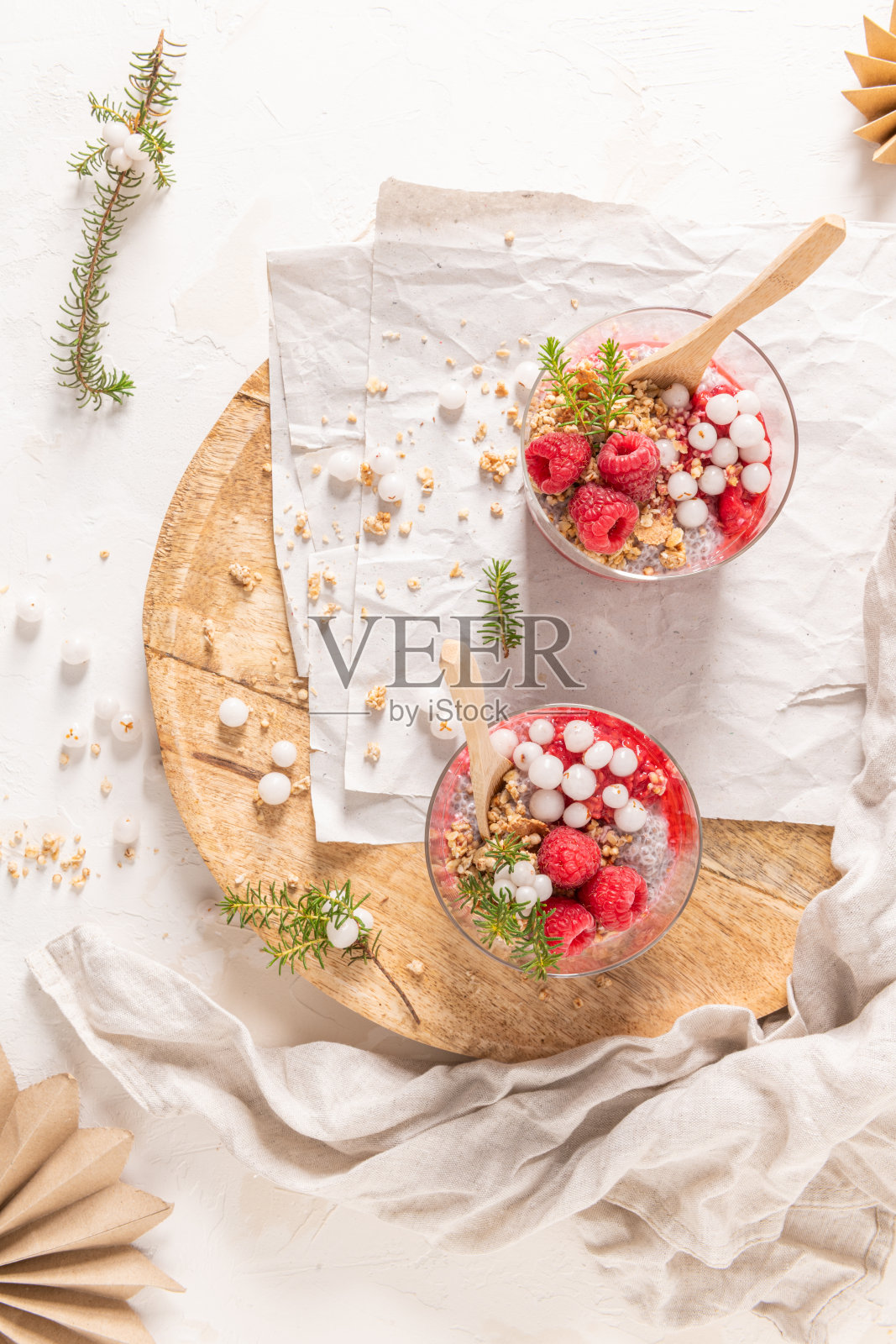 一张俯视图，展示了两杯分层的覆盆子和奇亚籽布丁，上面点缀着新鲜的覆盆子和白色越橘，摆放在一个质朴的木质托盘上。照片摄影图片