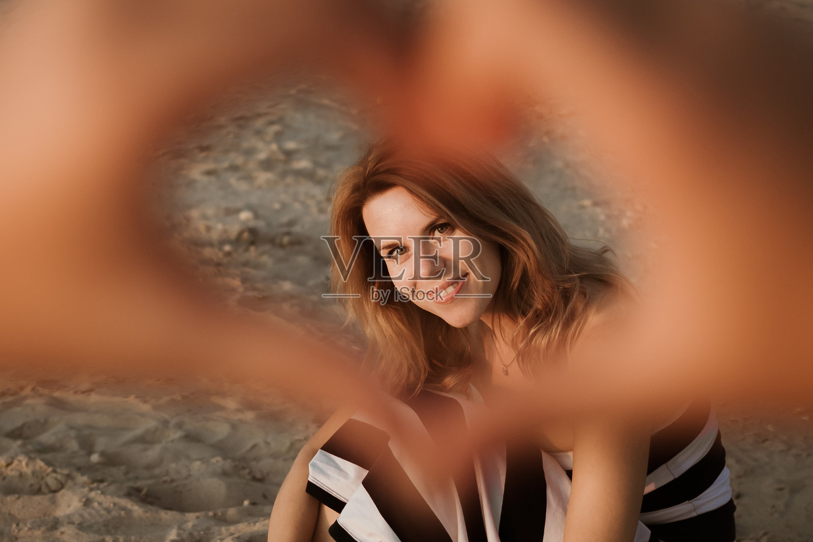 微笑的女人坐在海滩上，夕阳的余晖洒在身上，通过手心的心形手势看向镜头。爱的象征。父母与孩子之间的关系。母亲节。家庭的信任。幸福。照片摄影图片