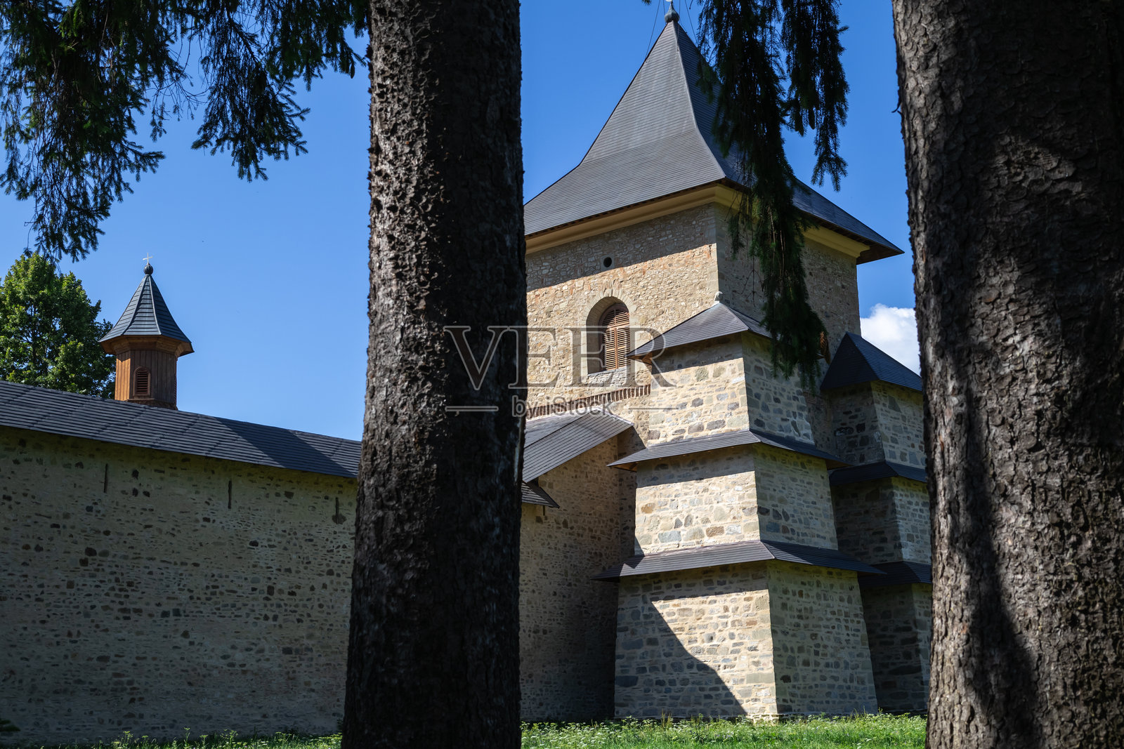 罗马尼亚的一座古老修道院，石墙和传统建筑，周围是绿草和高大的树木，蓝天清澈。照片摄影图片