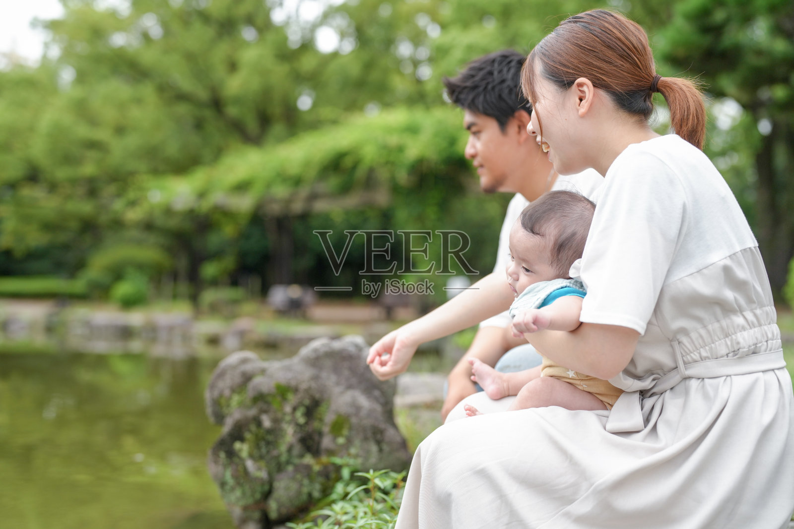 一家三口，一名男性和两名女性，坐在池塘边的一块岩石上。照片摄影图片