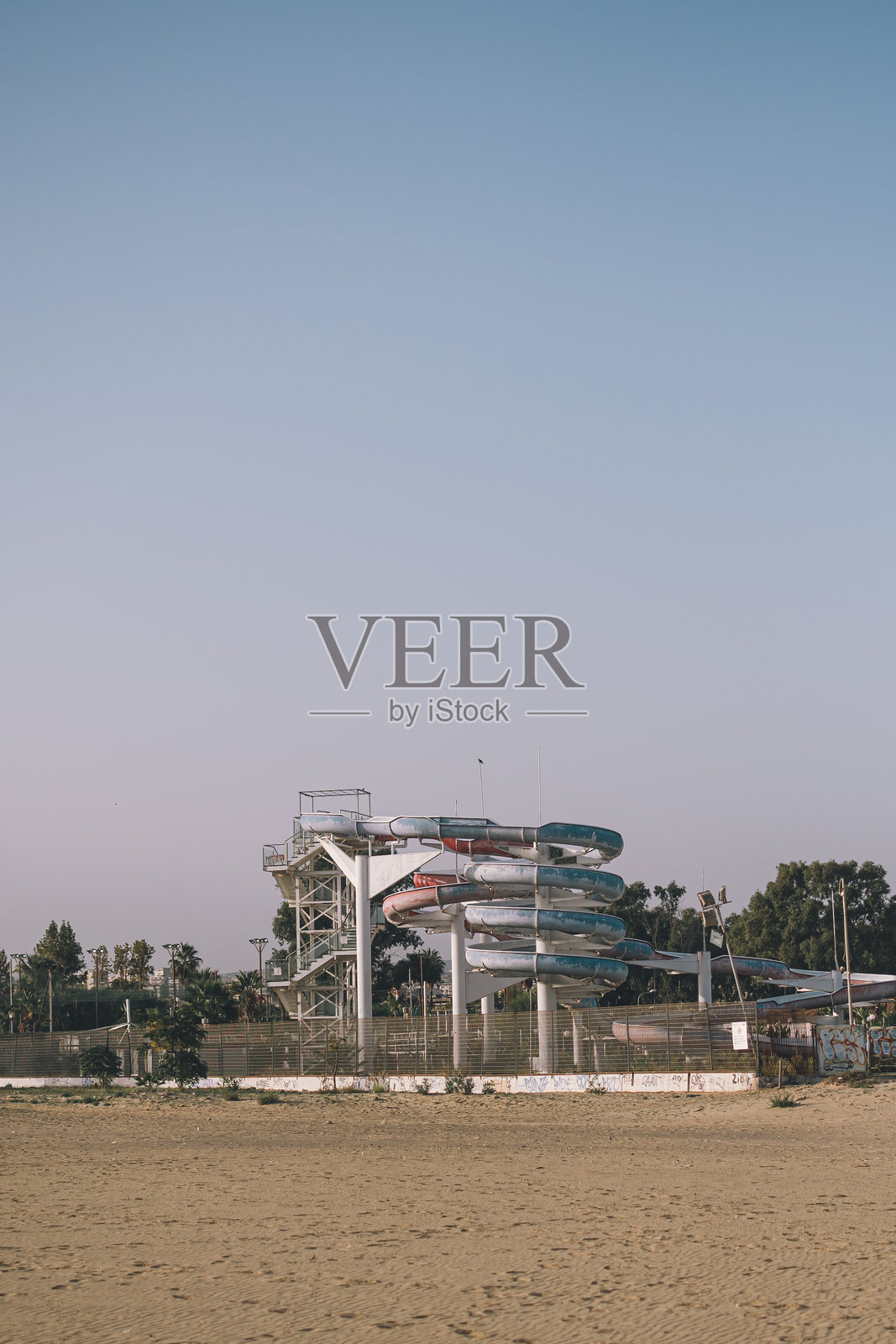 一个被遗弃的水上乐园，锈迹斑斑的五彩水滑梯在晴朗的蓝天下盘旋，周围是沙质地貌和郁郁葱葱的绿植，唤起对被遗忘的休闲场所的怀旧与魅力。意大利卡塔尼亚照片摄影图片